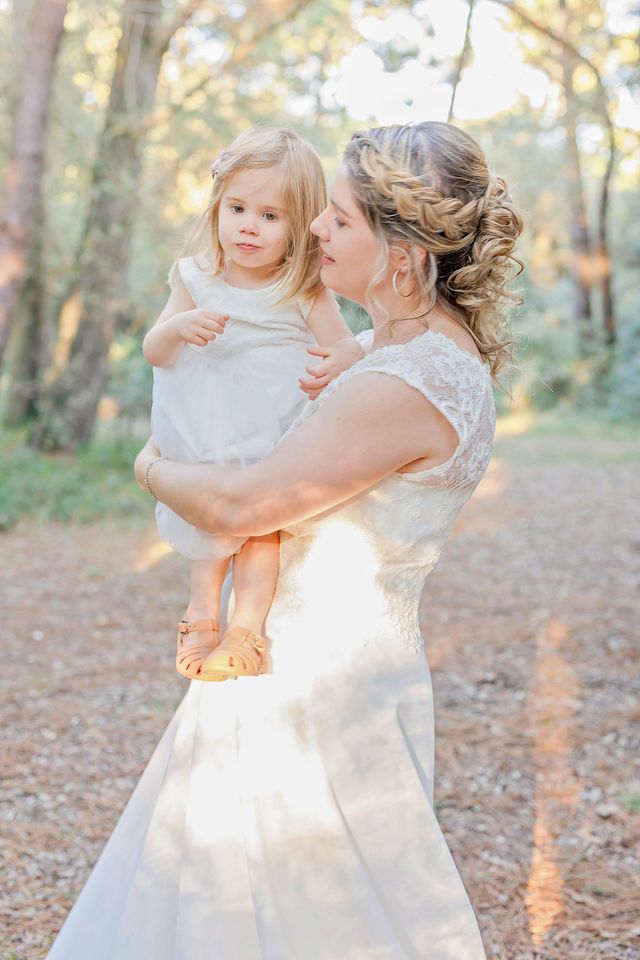 A joyful embrace between a bride and young child in a sunlit forest setting.
