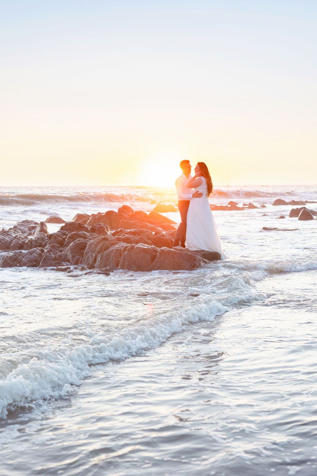 A couple in wedding attire stands on coastal rocks during a dramatic sunset.