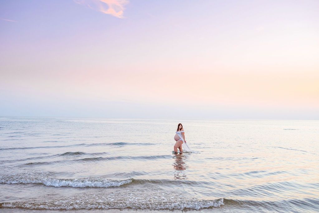 Woman in white dress walking through gentle ocean waves during sunset photoshoot.