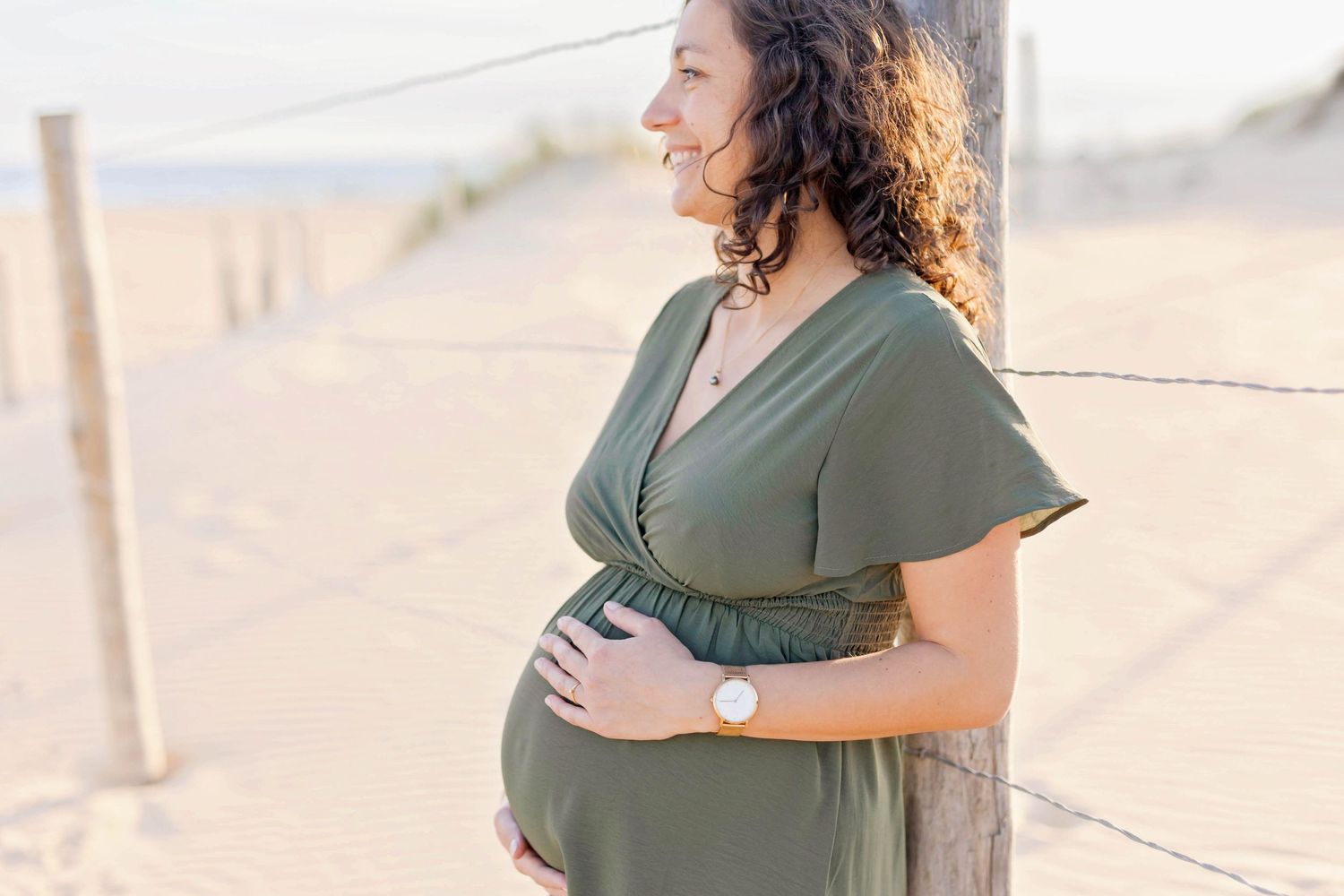 A woman in olive dress cradles her baby bump while standing on a sunny beach.