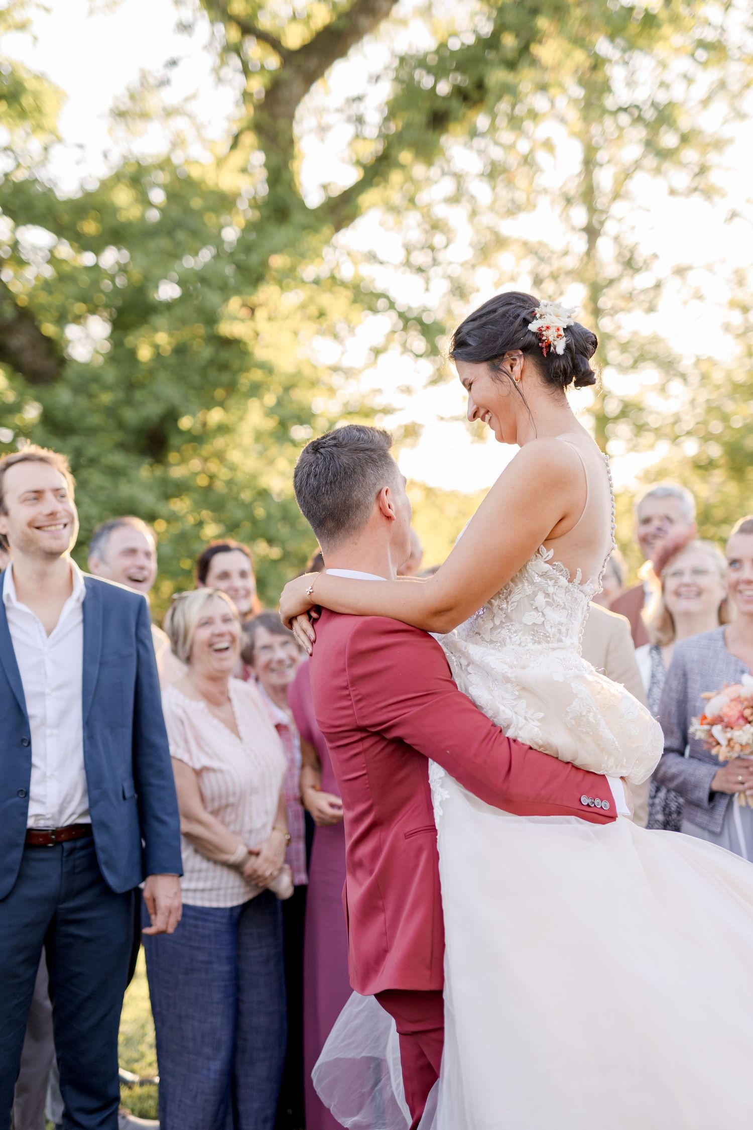 Wedding couple shares intimate moment as groom lifts bride while guests watch and smile in garden setting.