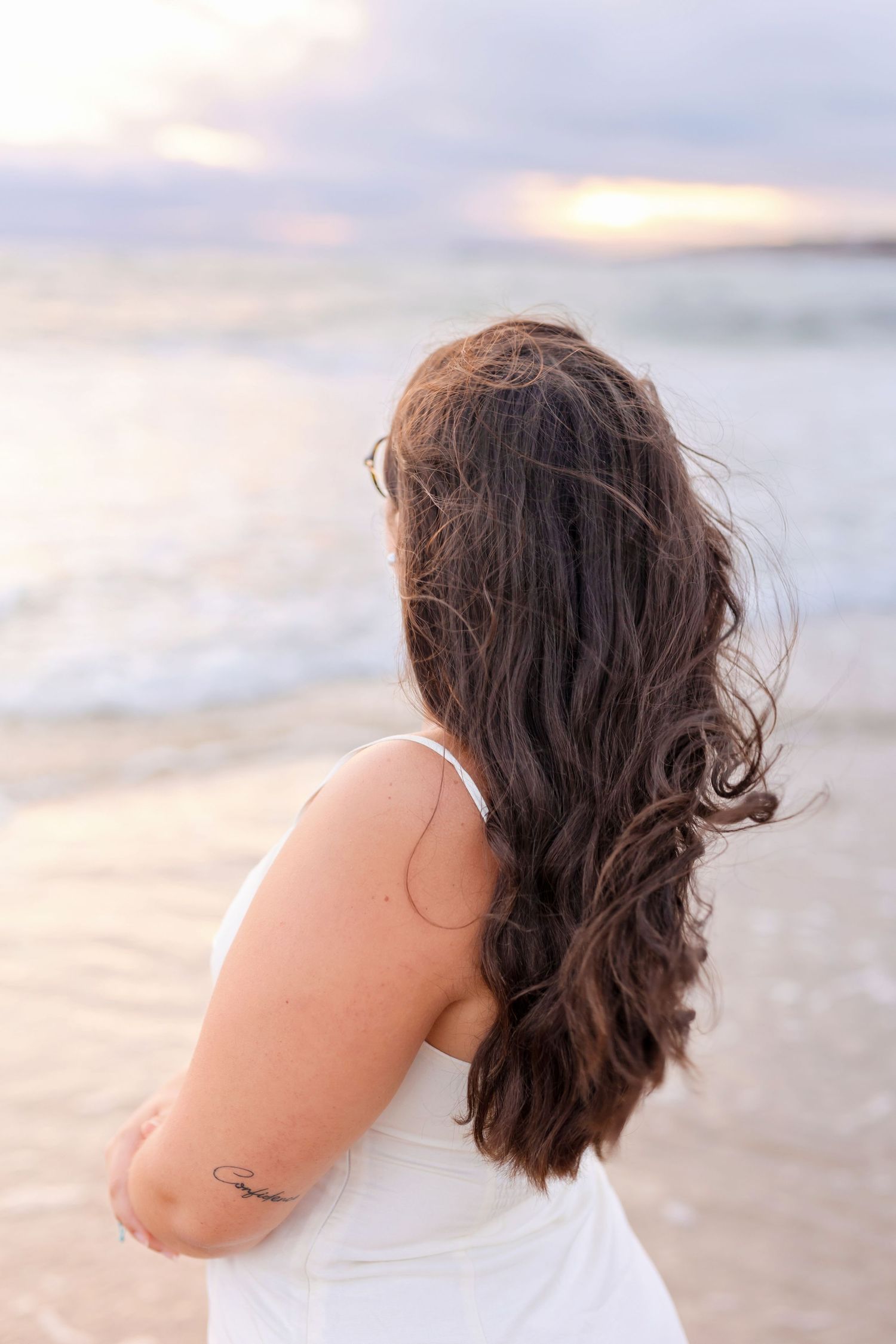 Back view of person with long dark wavy hair wearing white at sunset on beach.