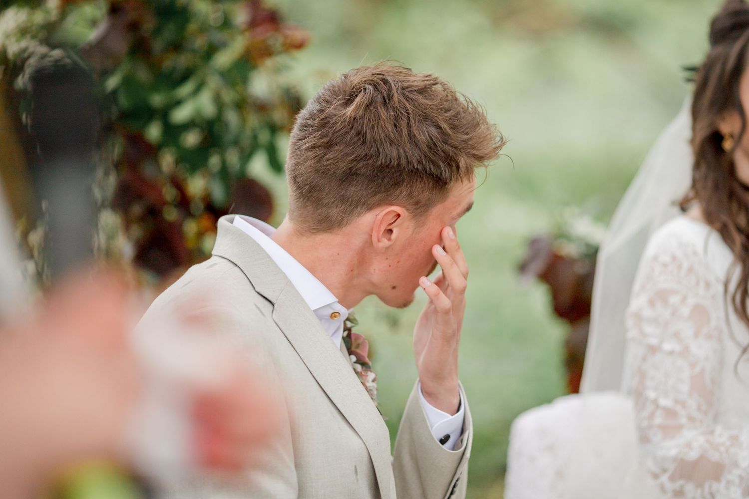 Guest in light colored suit wipes tears during emotional outdoor ceremony moment.