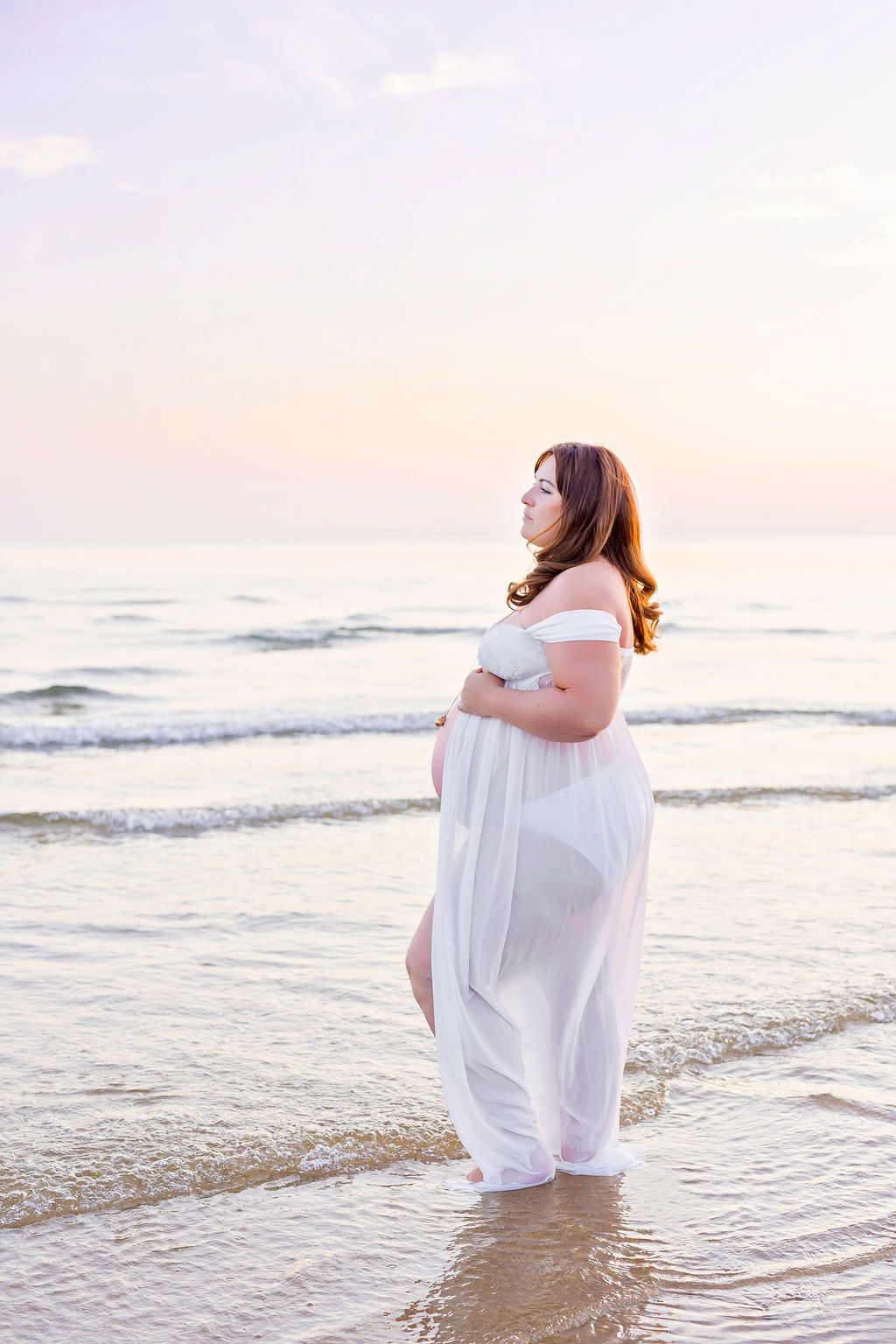 Expectant woman in flowing white dress stands peacefully at the ocean's edge during sunset.