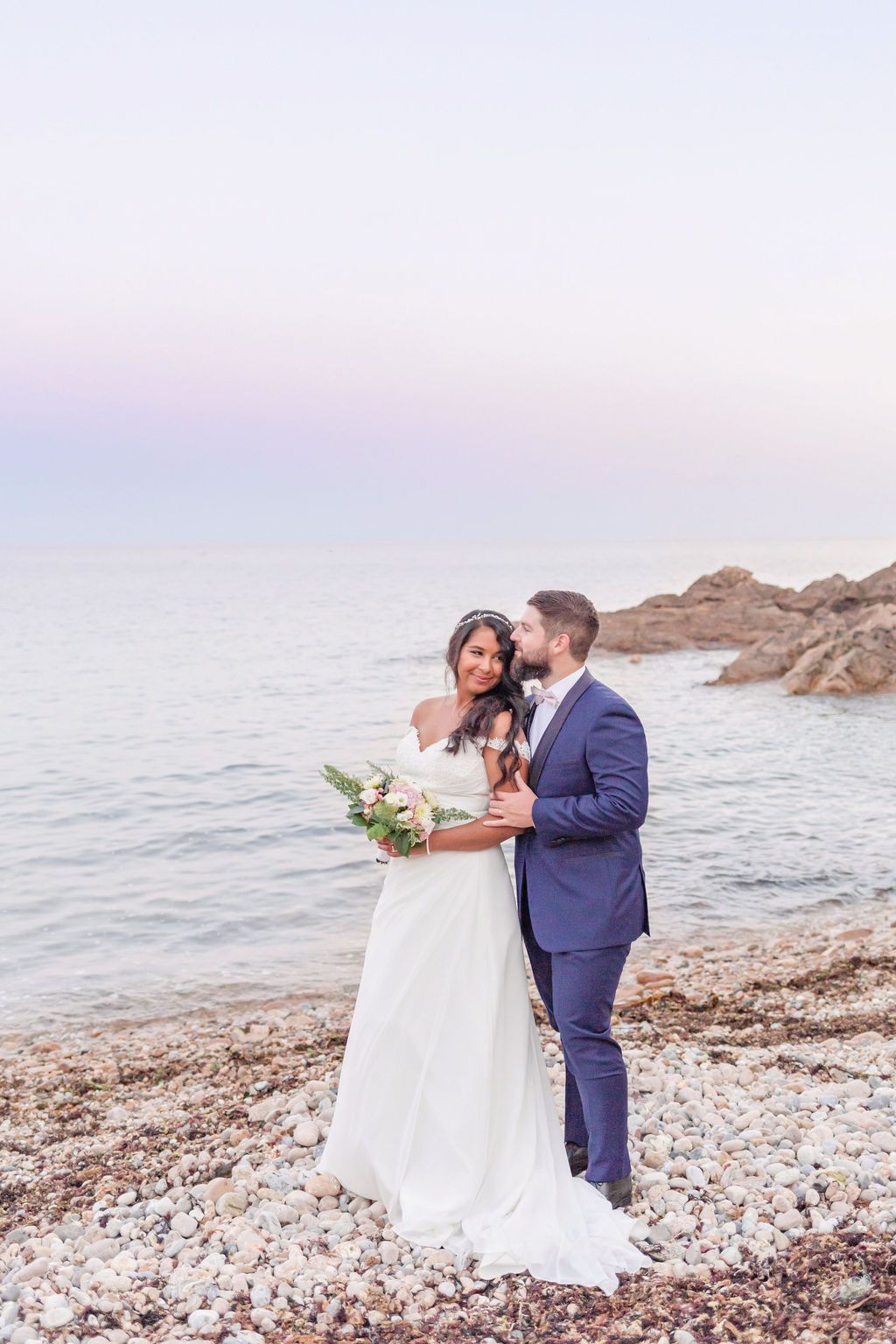 Elegant coastal wedding portrait with bride holding bouquet against pastel sunset sky.