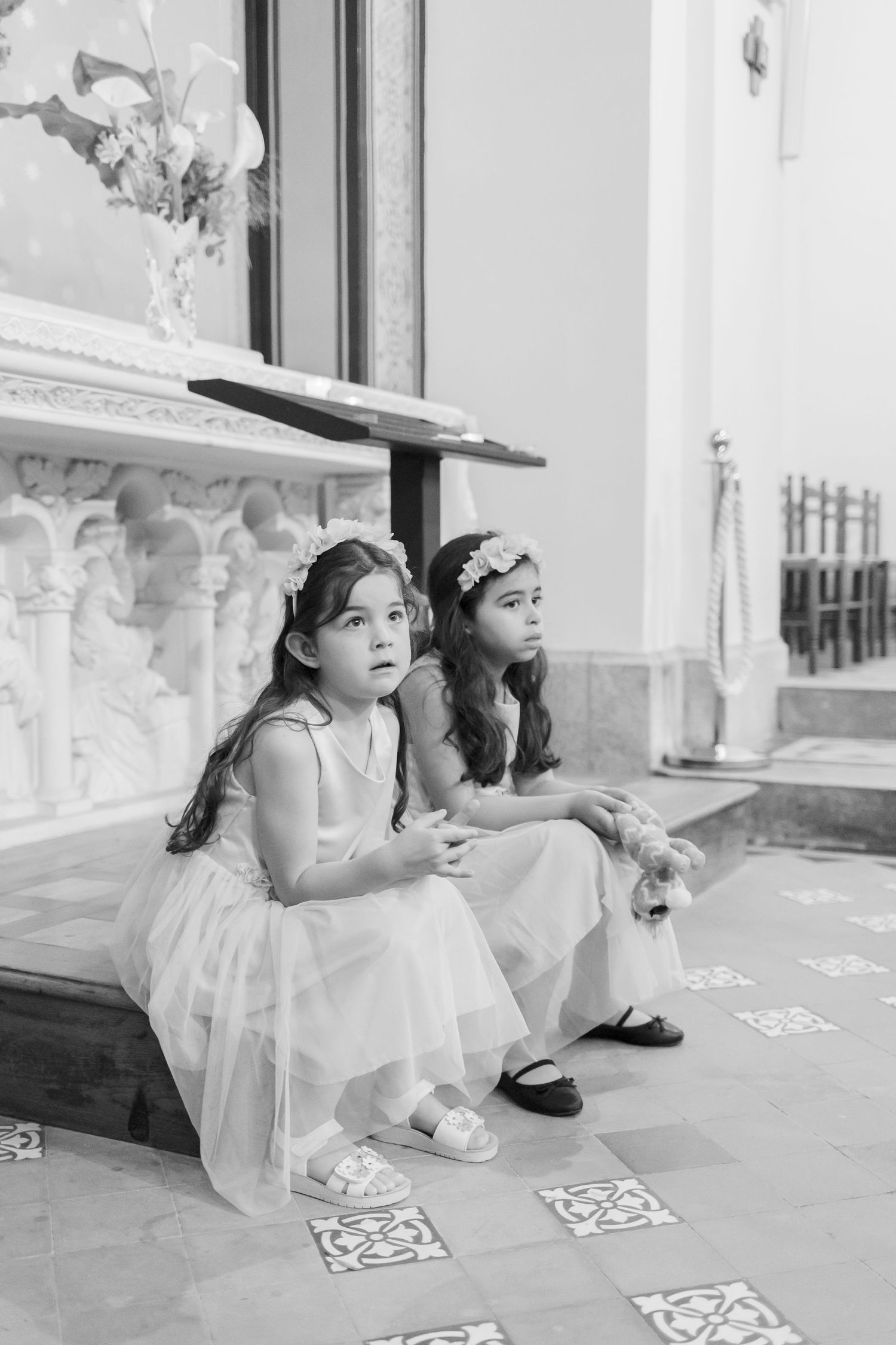 Two young flower girls in white dresses sit on church steps during a wedding ceremony.