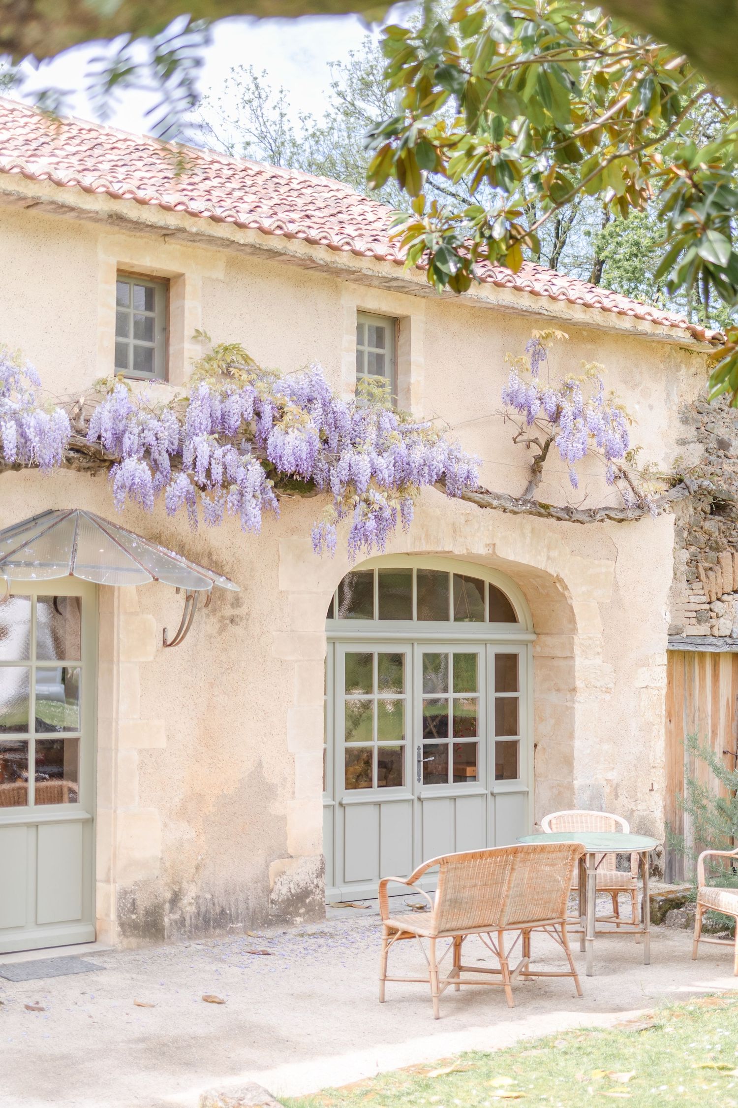 Charming cottage exterior with blooming wisteria and rustic furniture on a patio beneath an arched doorway.