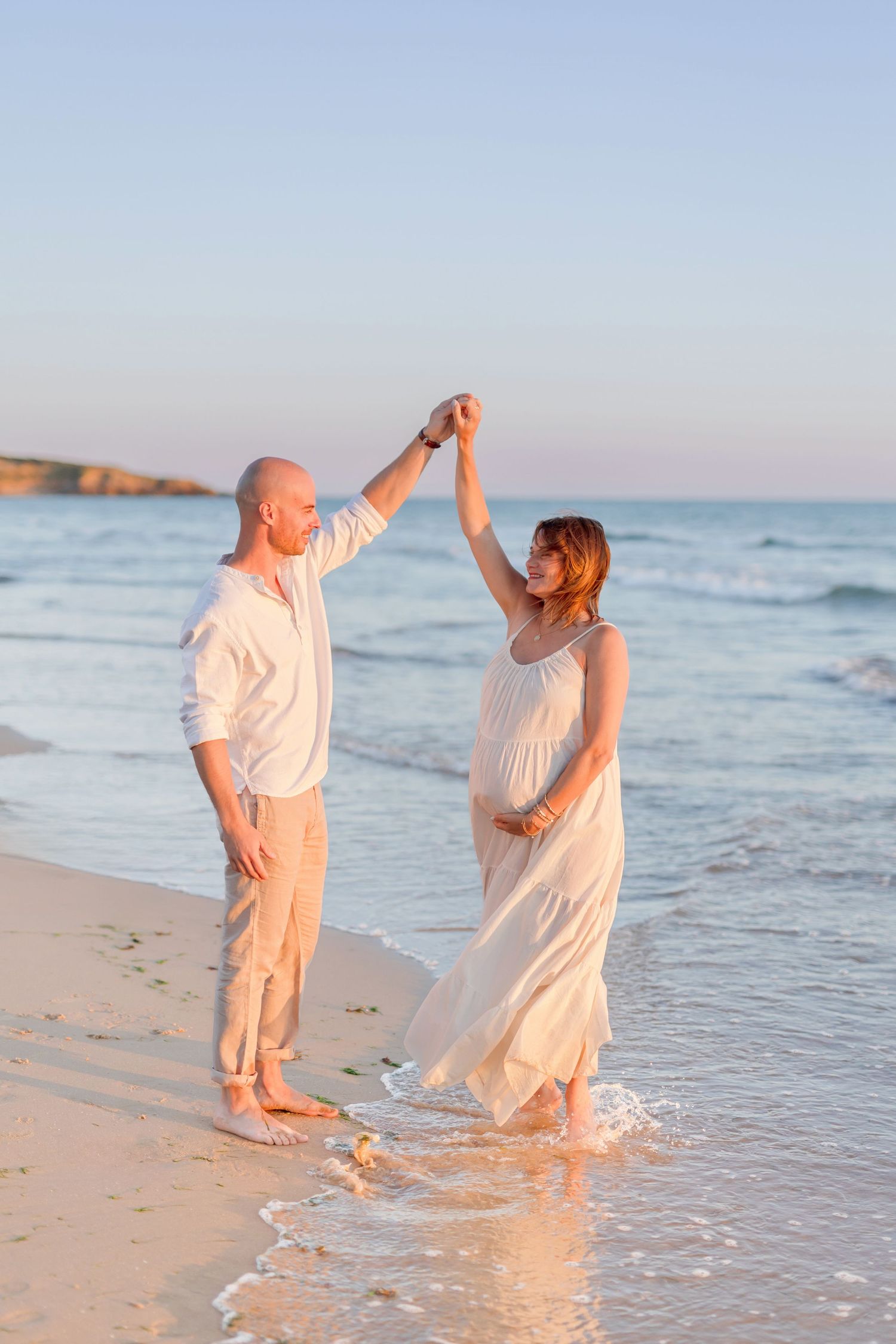 Dancing couple in white outfits enjoying sunset at the beach with waves in background.
