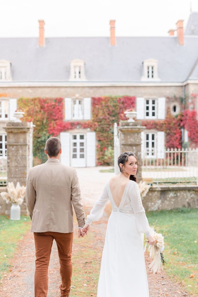 A couple in wedding attire walks along a stone walkway near a classical French chateau with autumn foliage.