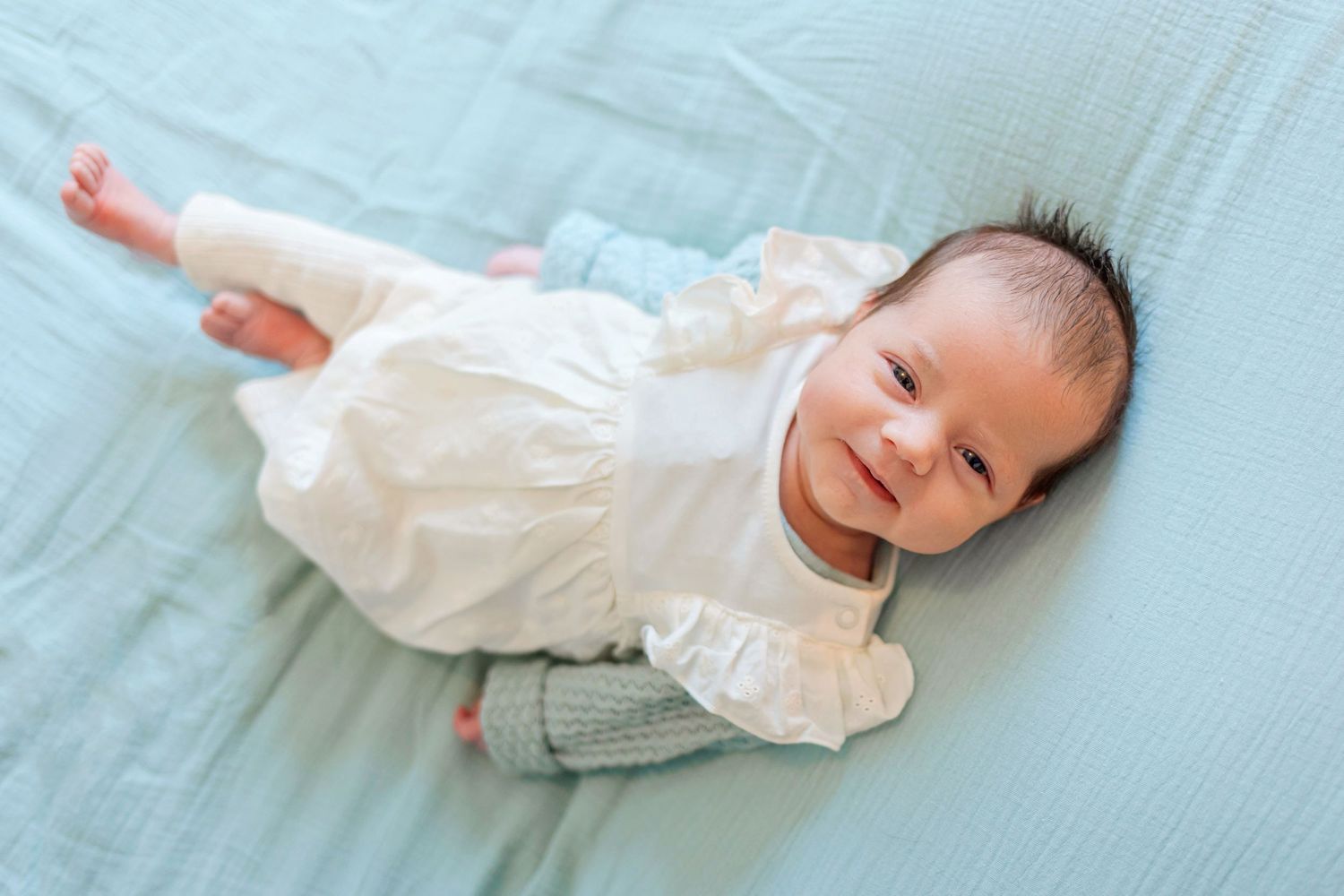 Smiling newborn baby wearing white outfit lies on light blue bedding.