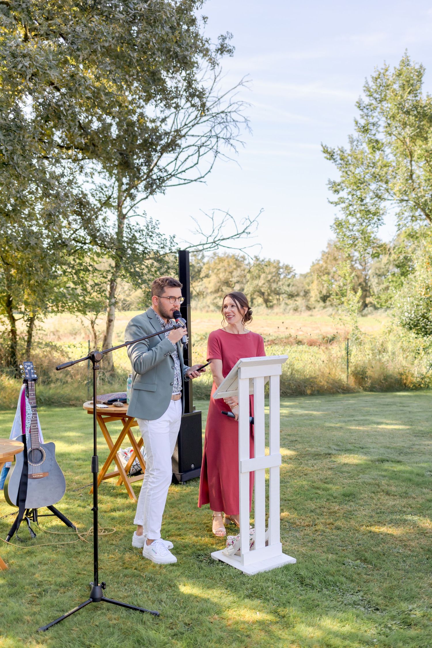 Outdoor wedding ceremony with speakers at white podium surrounded by natural scenery.