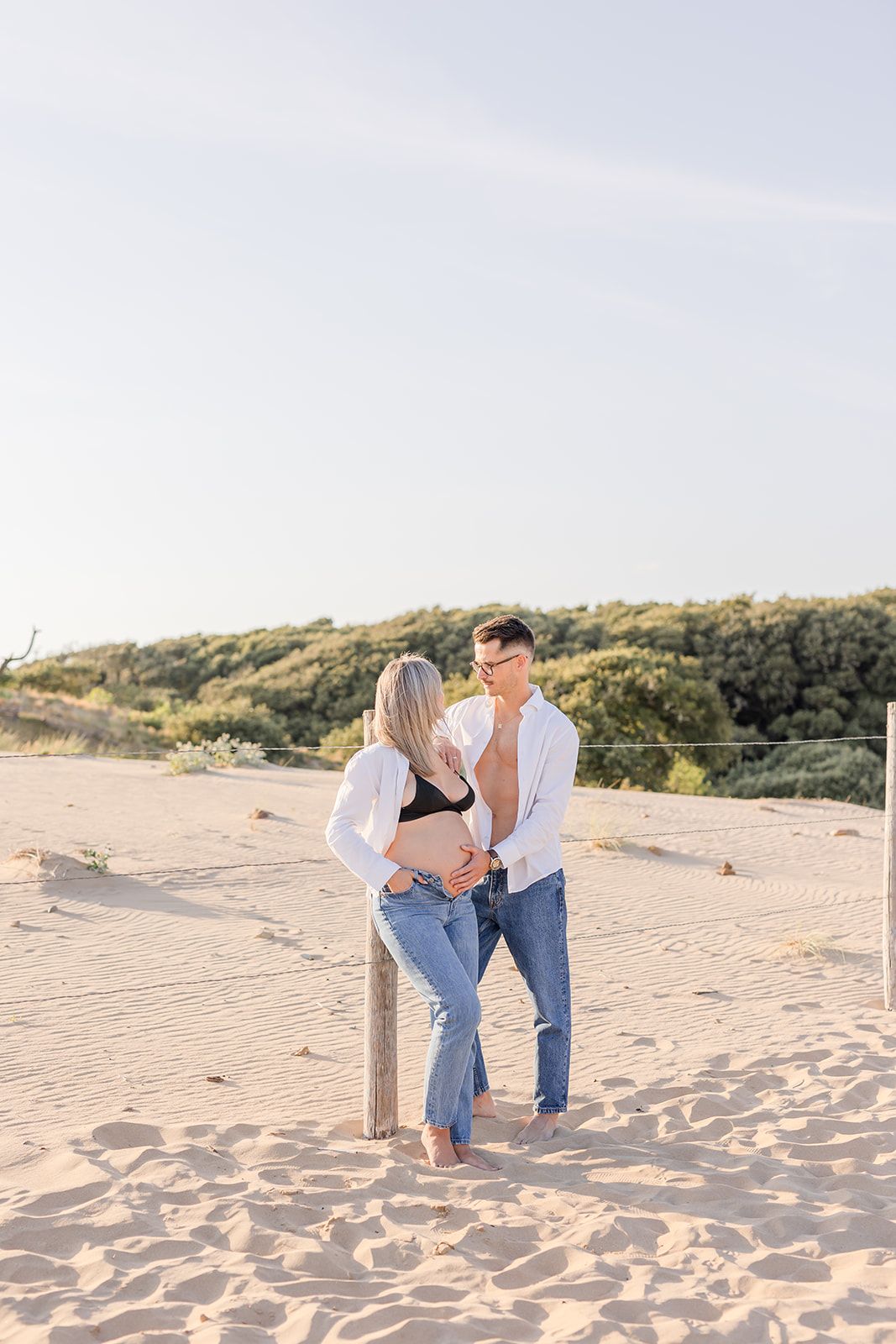 A couple in light colored clothing share an intimate moment while standing together on sandy beach dunes at sunset.