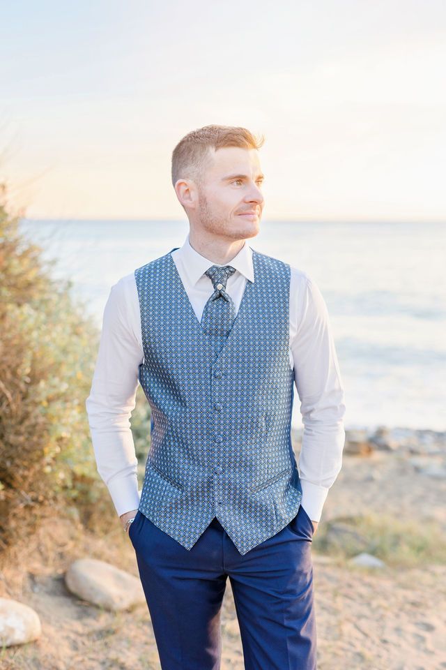 Groom in formal attire with blue vest poses on beach at sunset.