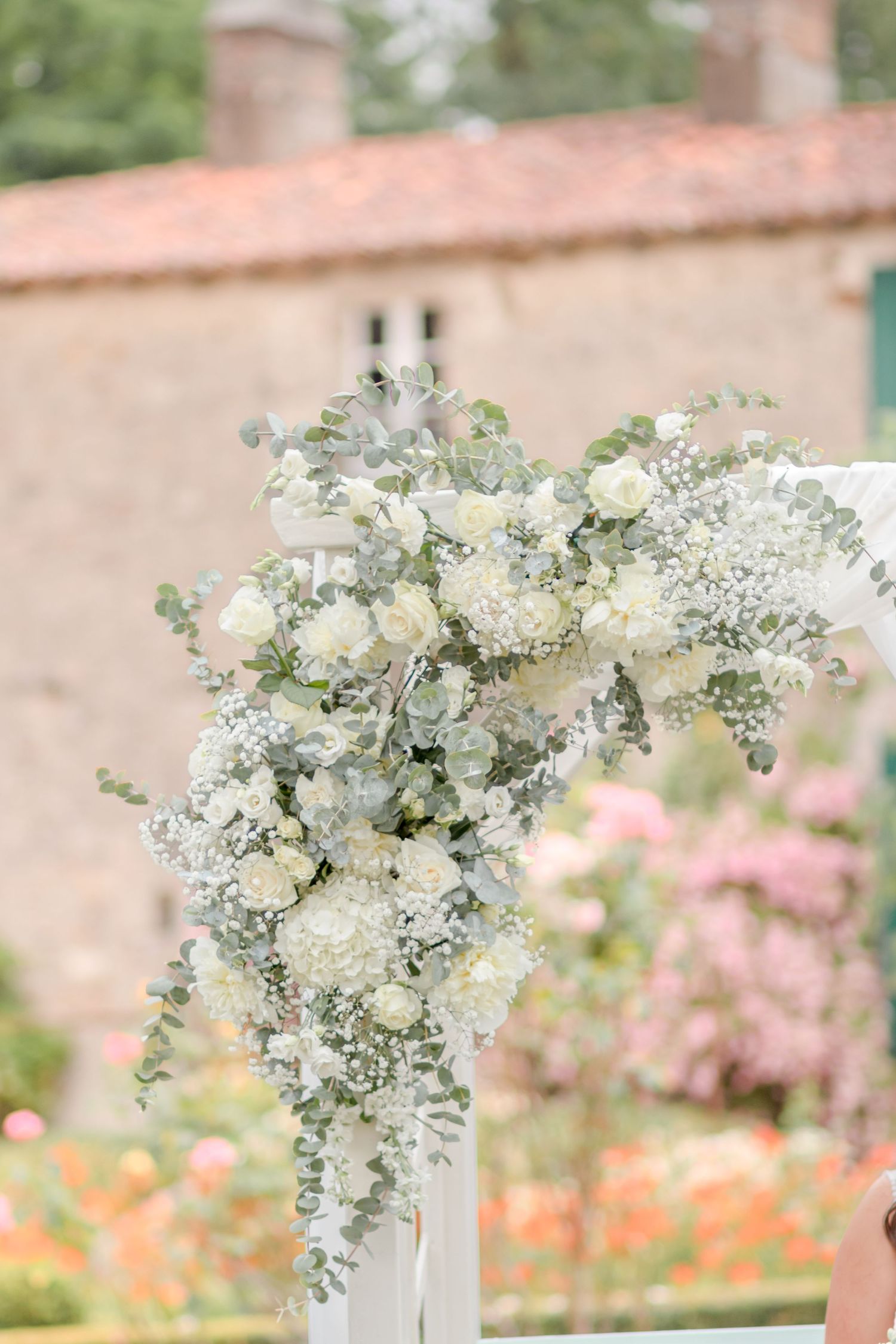 Elegant white floral arrangement with eucalyptus on a wedding arch against a pink stone building.