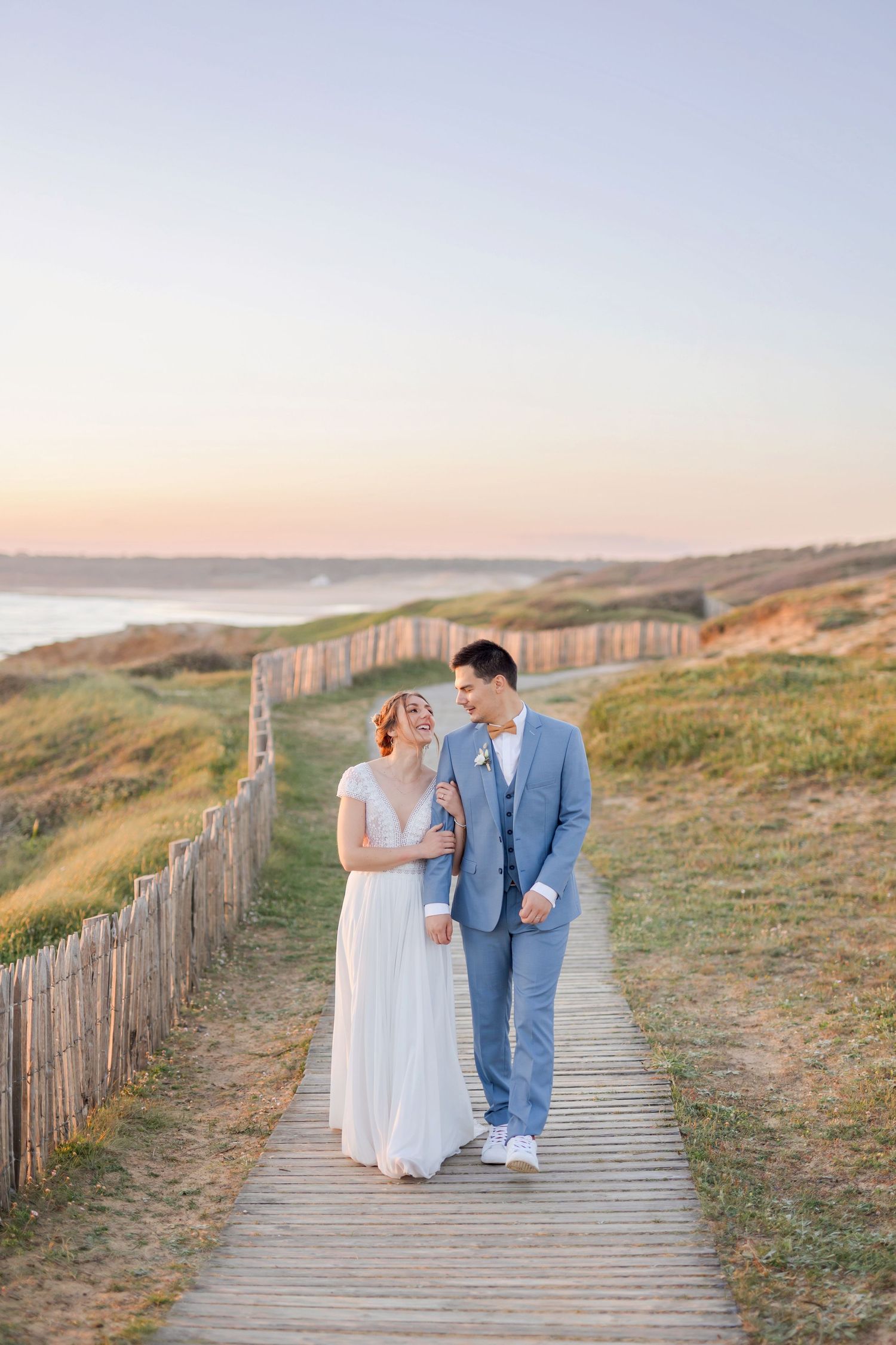 Couple in wedding attire strolling together on seaside boardwalk during sunset.