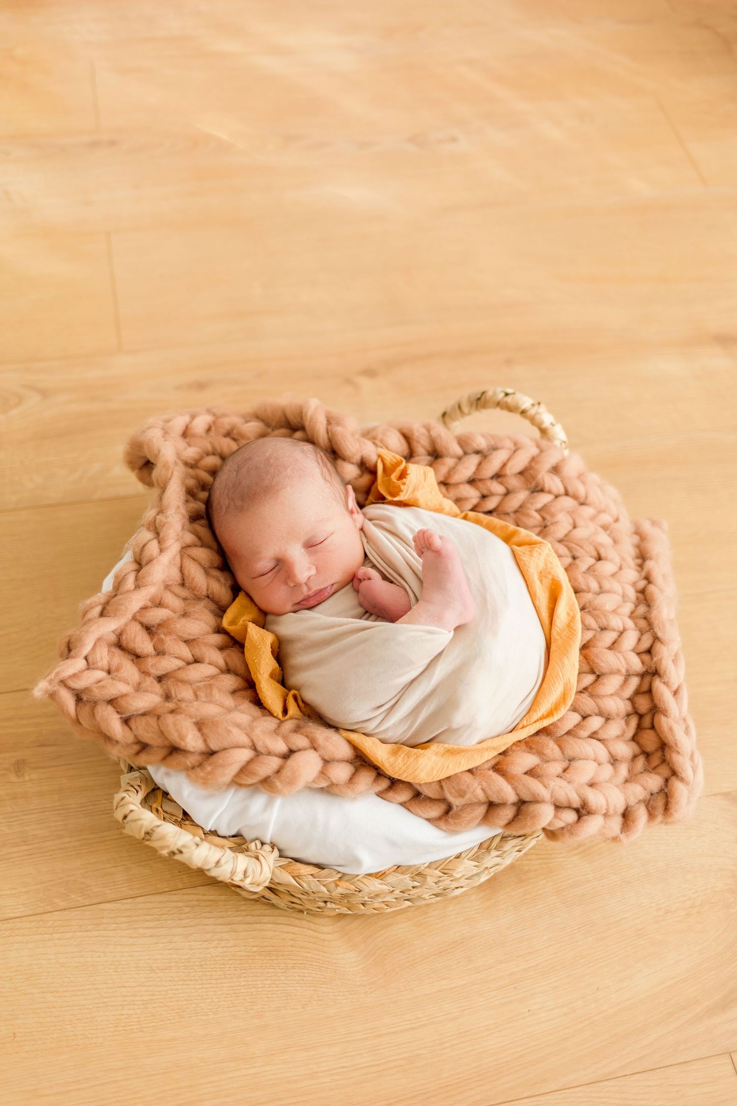 Overhead view of newborn baby sleeping on beige knitted blanket in woven basket.