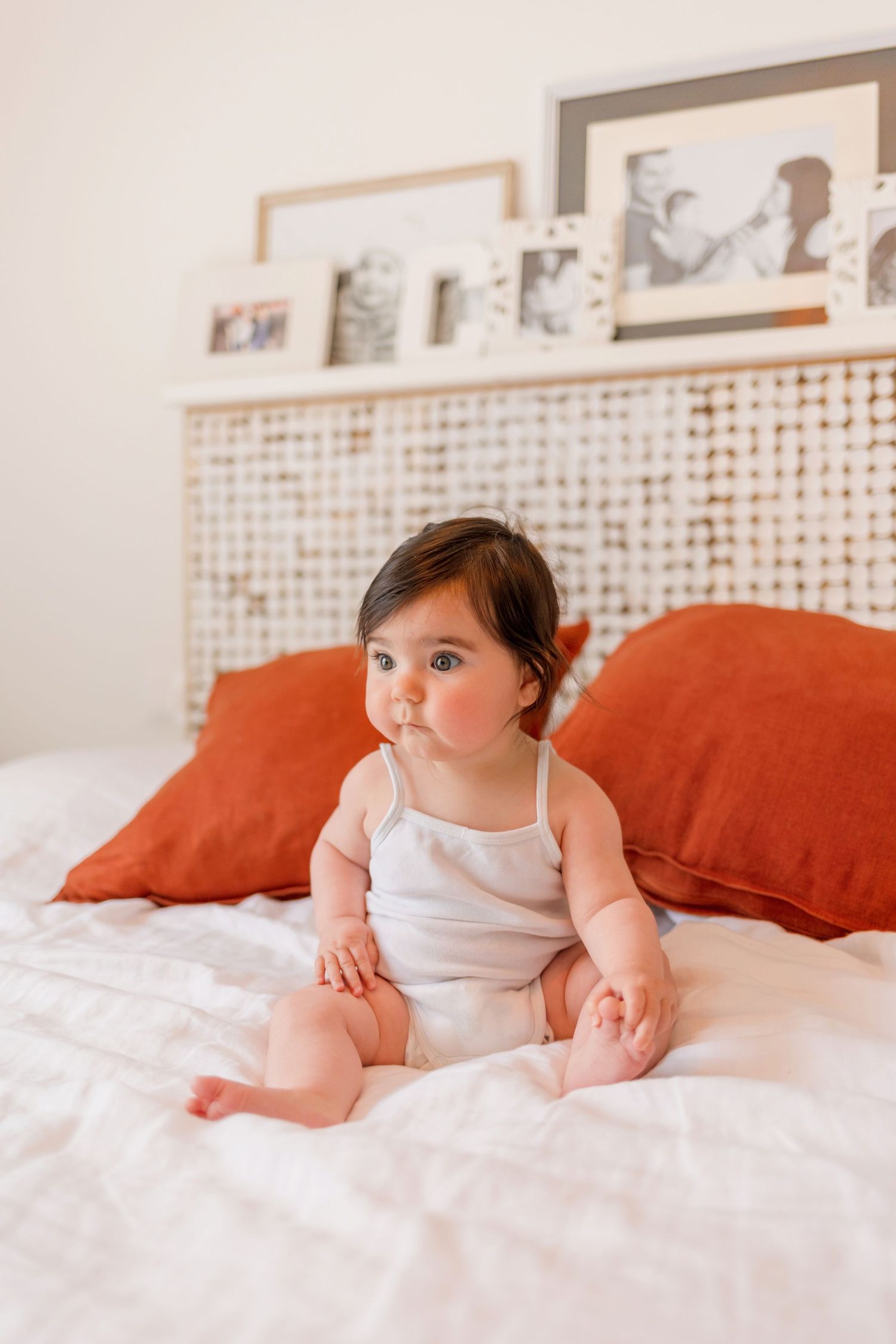Baby in white onesie sitting on white bedding with orange throw pillows.