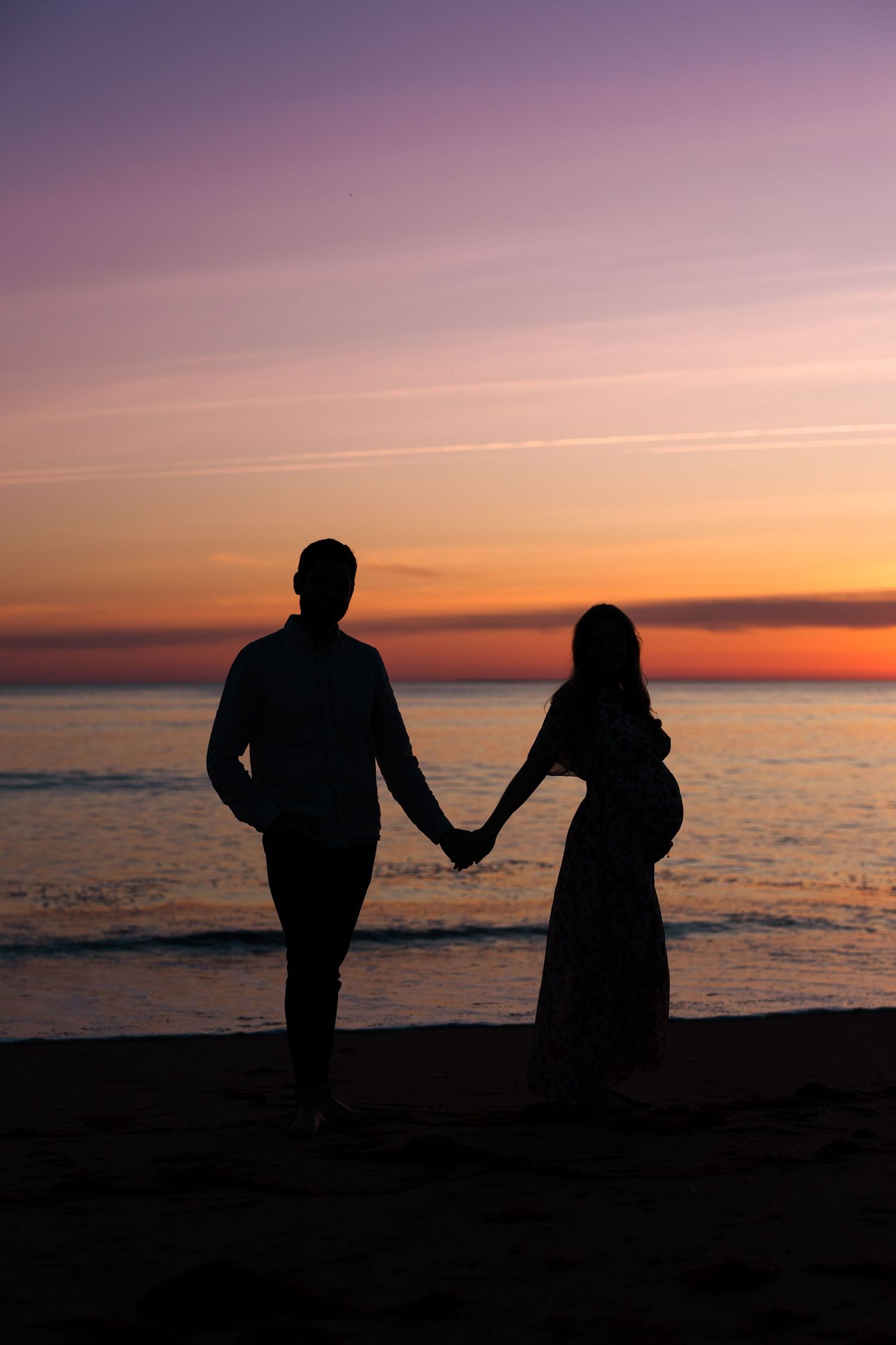 Silhouetted couple holding hands walking along beach at dramatic purple sunset.