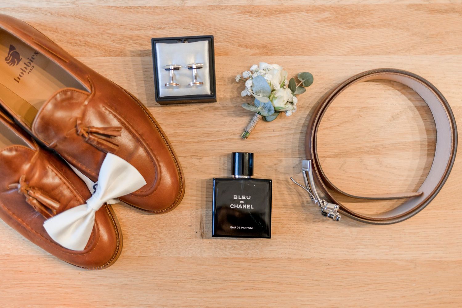 Wedding accessories including brown leather shoes and belt arranged on wooden surface.