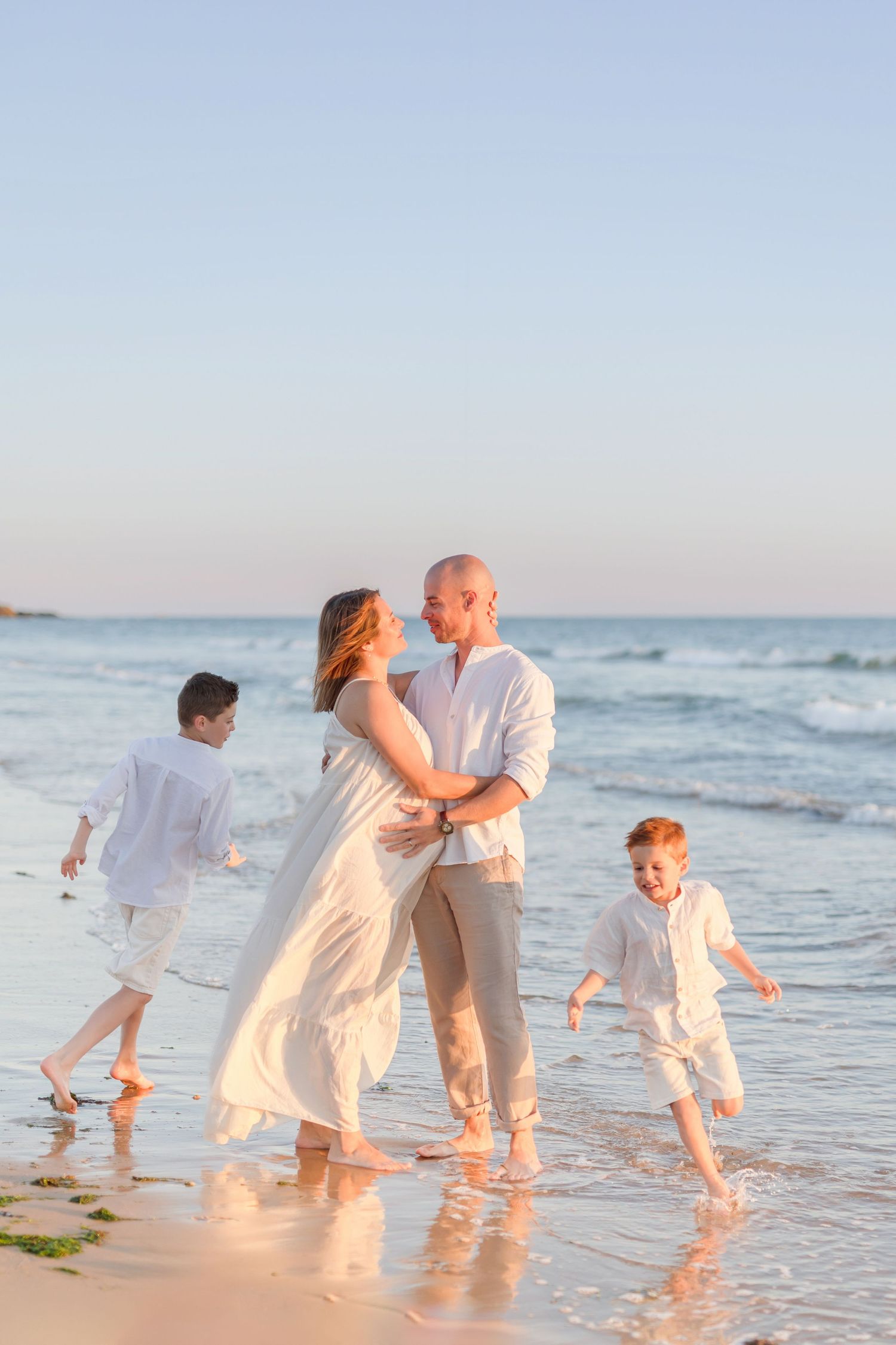 Family playing and running along beach shoreline in white outfits at sunset.