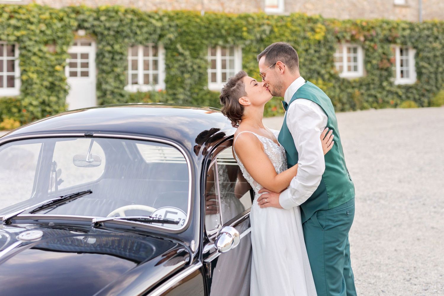 Wedding couple poses romantically beside a vintage black Volkswagen Beetle against ivy-covered wall.