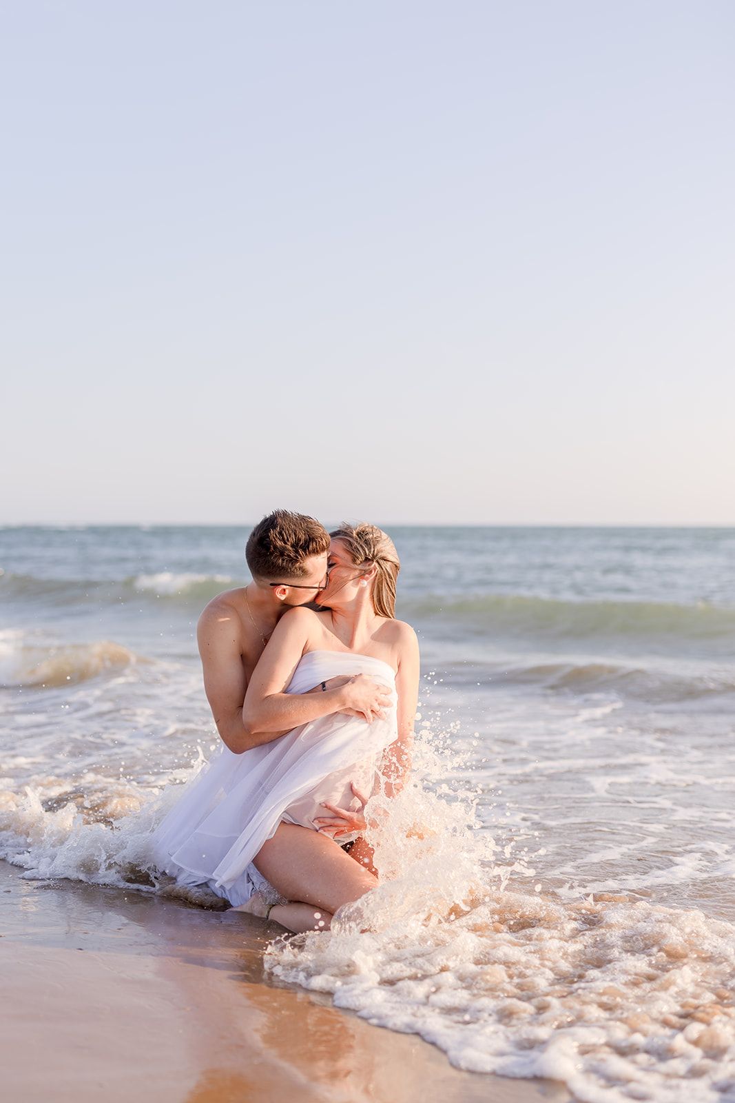 A romantic couple sitting in white outfits gets splashed by ocean waves on a sandy beach at sunset.