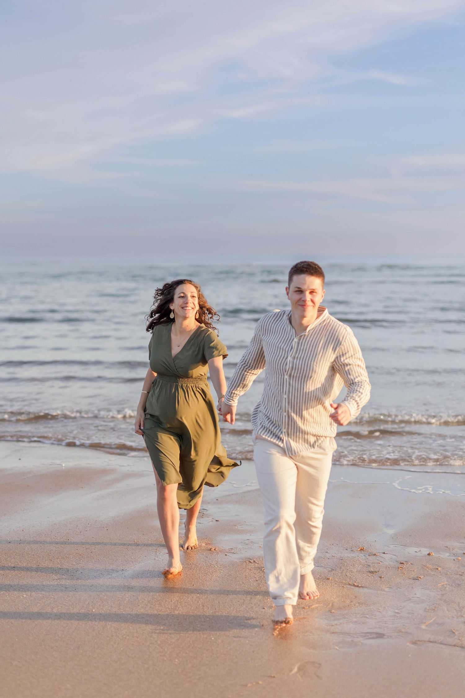 A couple runs joyfully along a sandy beach with waves crashing behind them in warm sunlight.