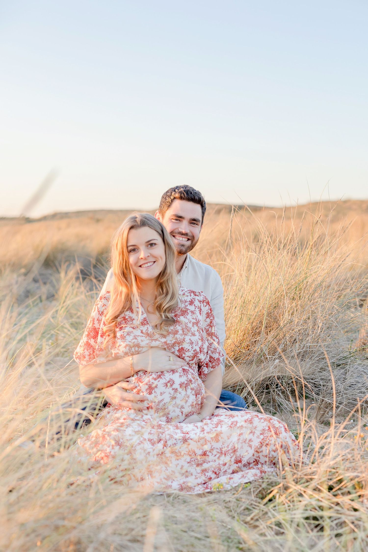 A couple sits together in a golden field of tall grass during sunset with the person in a floral dress.
