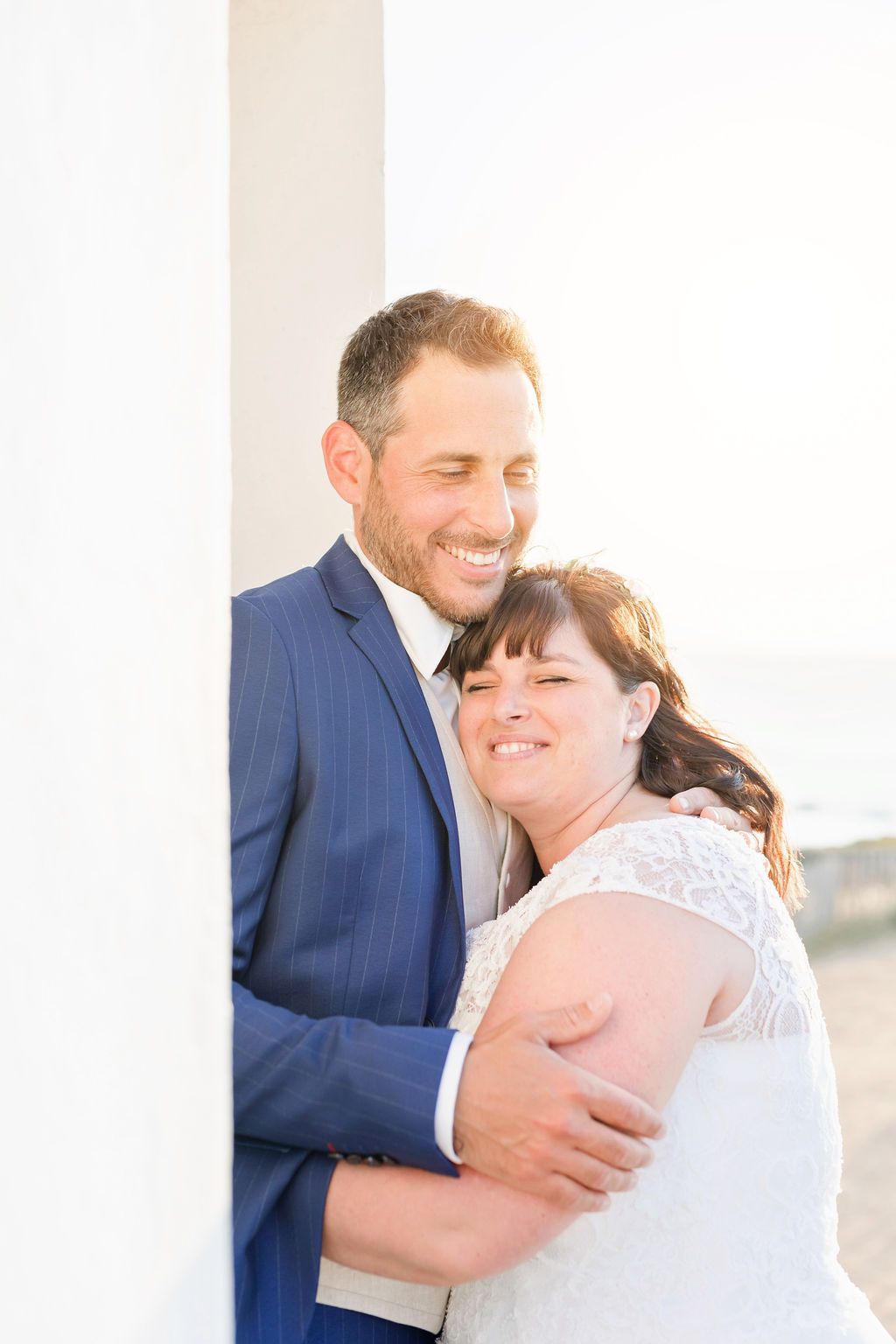 A tender moment between newlyweds embracing against a white lighthouse backdrop.