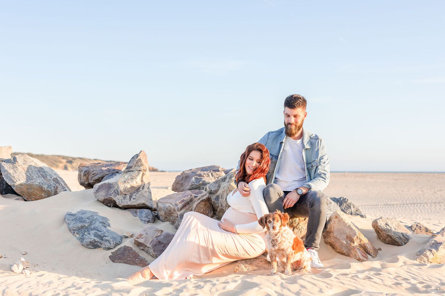 Couple relaxes with dog among beach rocks during golden hour maternity shoot.