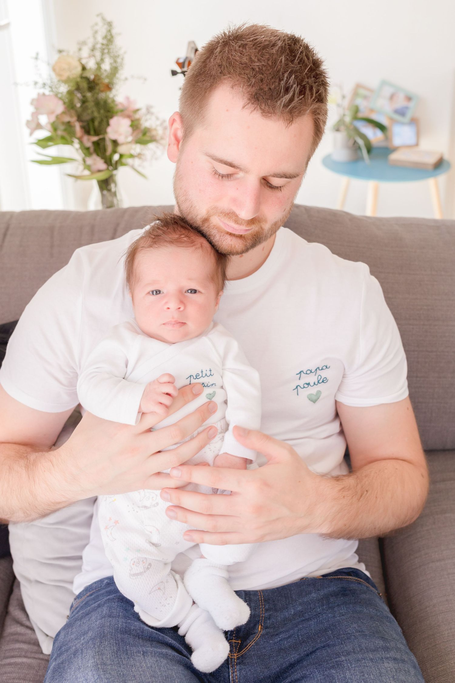Parent in white t-shirt holding sleeping baby in white onesie on living room couch.