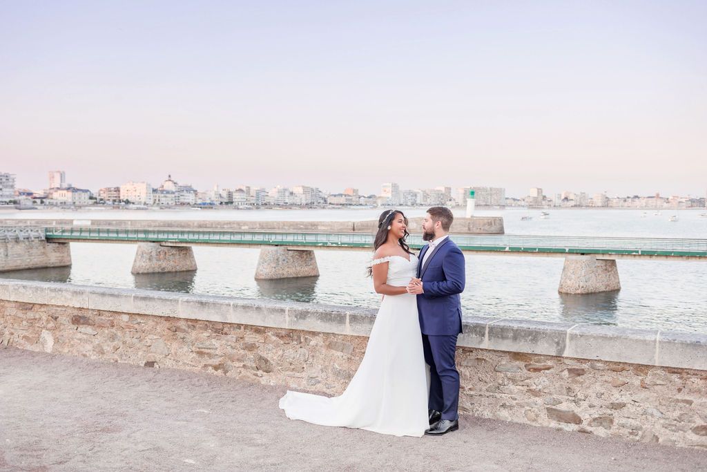 A couple in wedding attire embraces on a stone walkway overlooking a coastal cityscape at sunset.