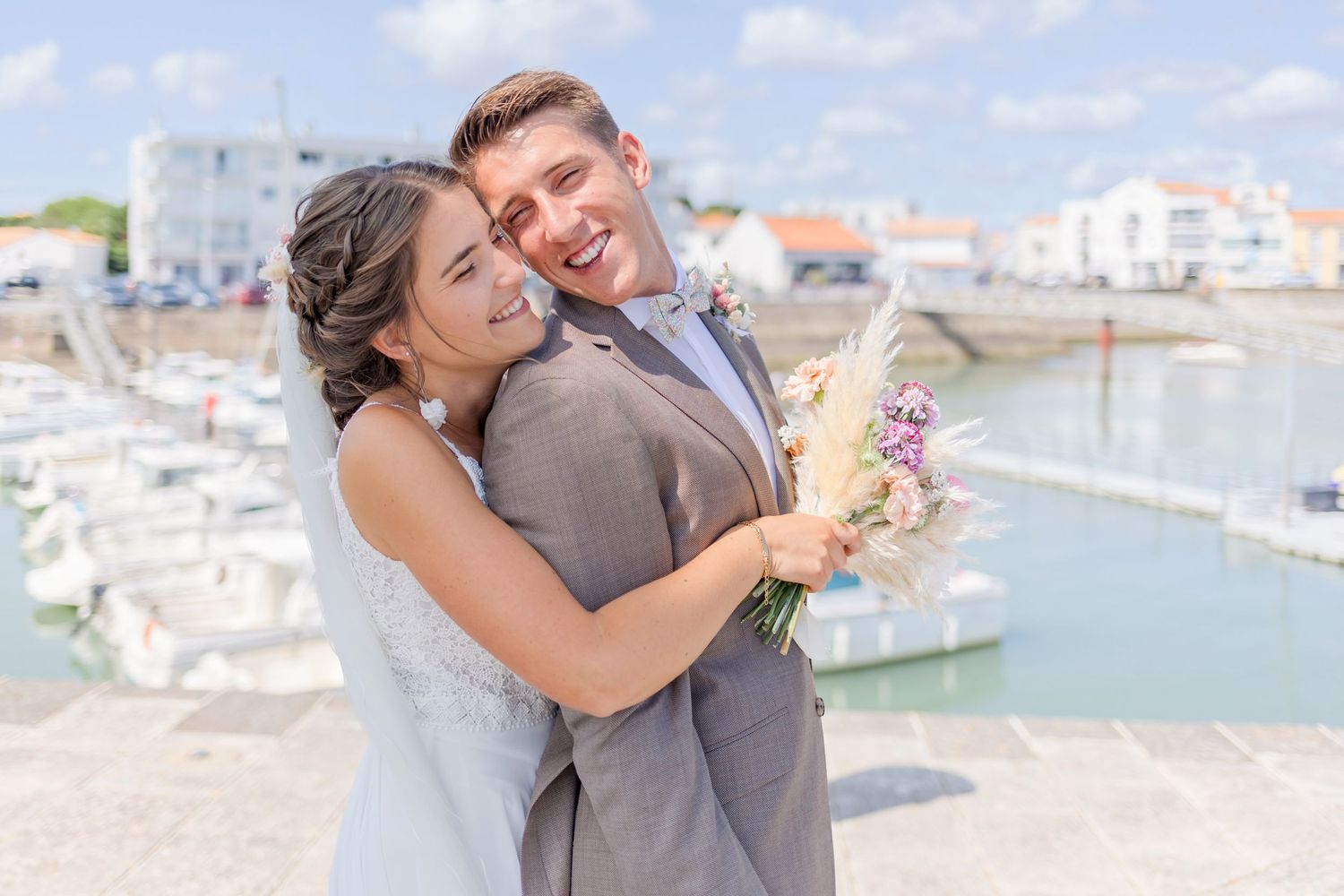 Joyful wedding couple sharing laughs near harbor with boats in background.