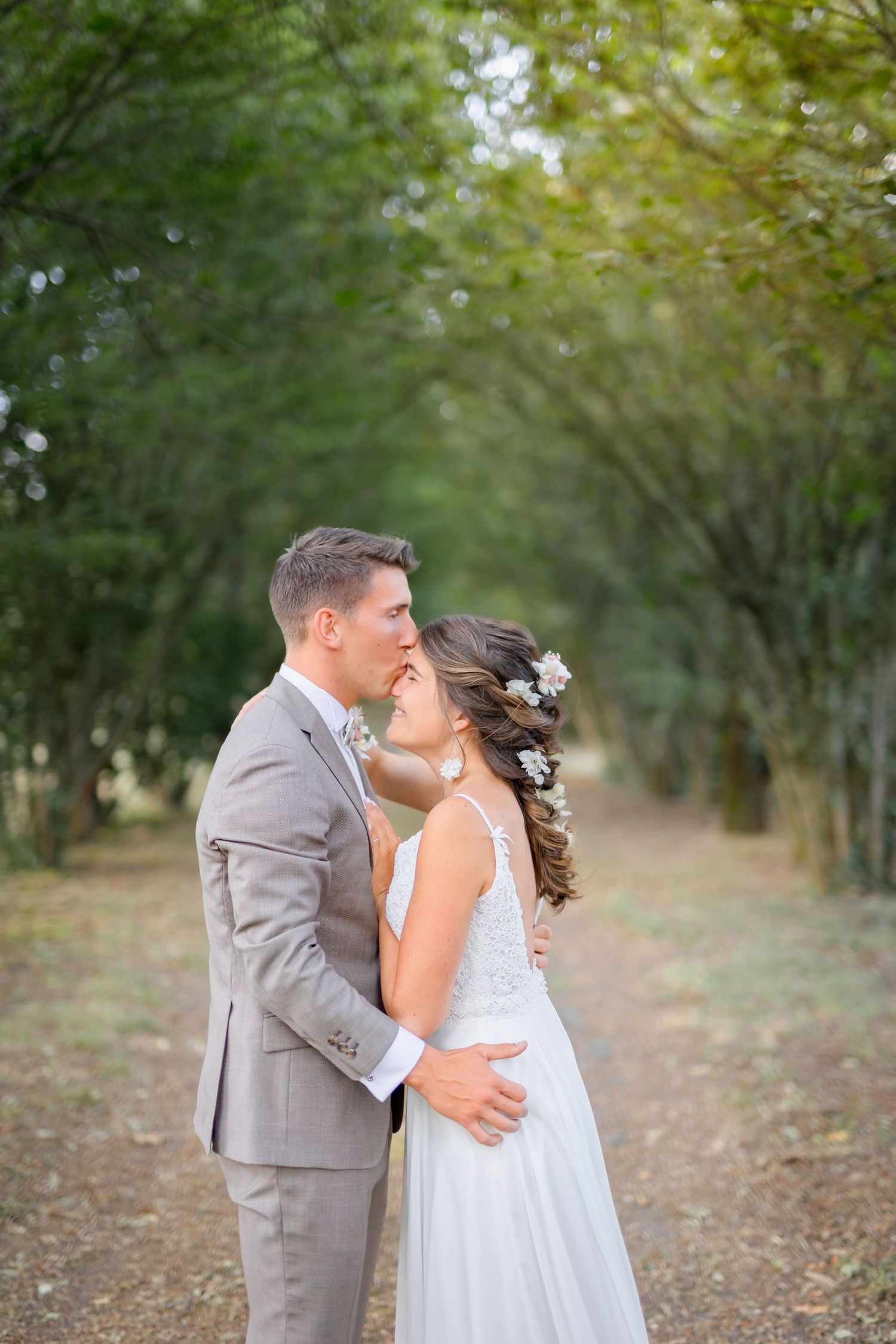 Romantic embrace between newlyweds on a tree-lined path with soft afternoon light.