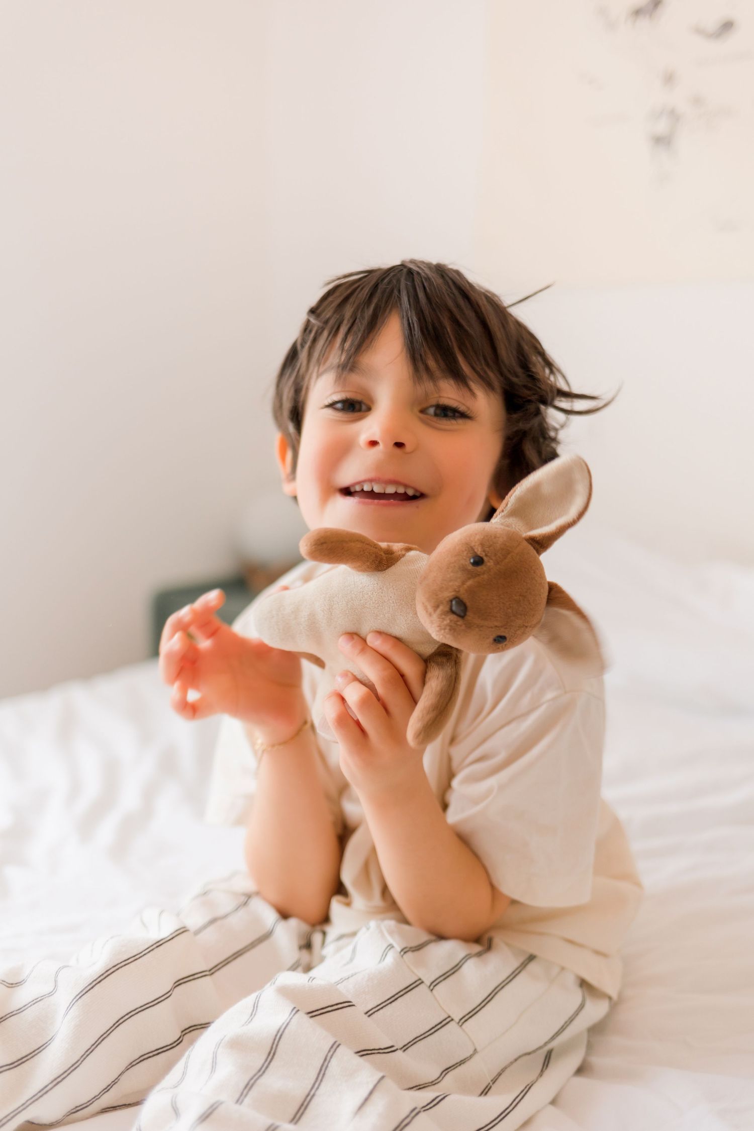 A child in white plays joyfully with a stuffed bunny toy on a white bed.
