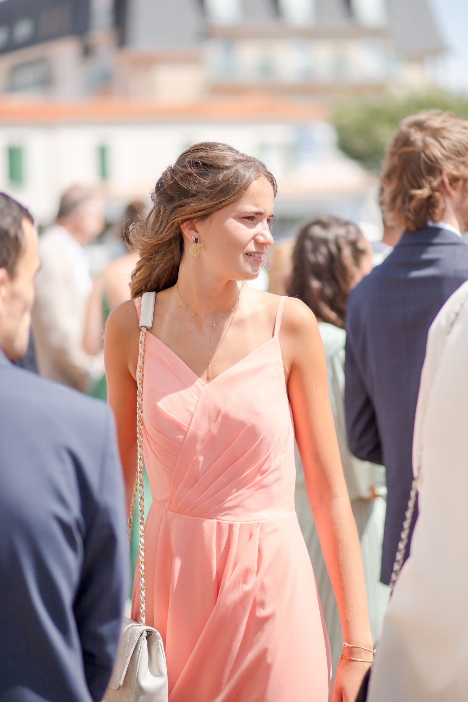 Person in coral pink dress walking outdoors among crowd wearing formal attire.