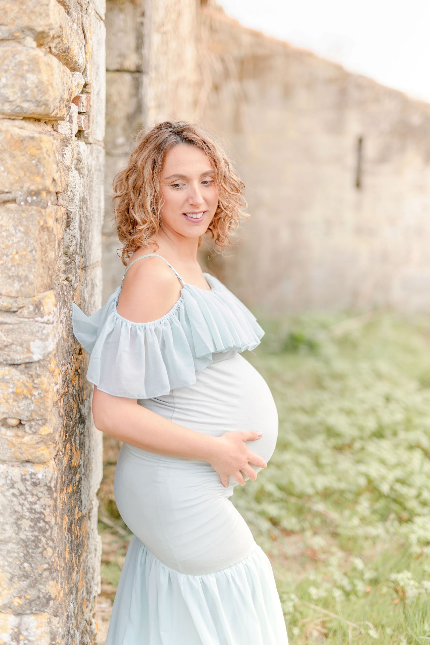 Pregnant woman in blue dress poses against rustic stone wall.