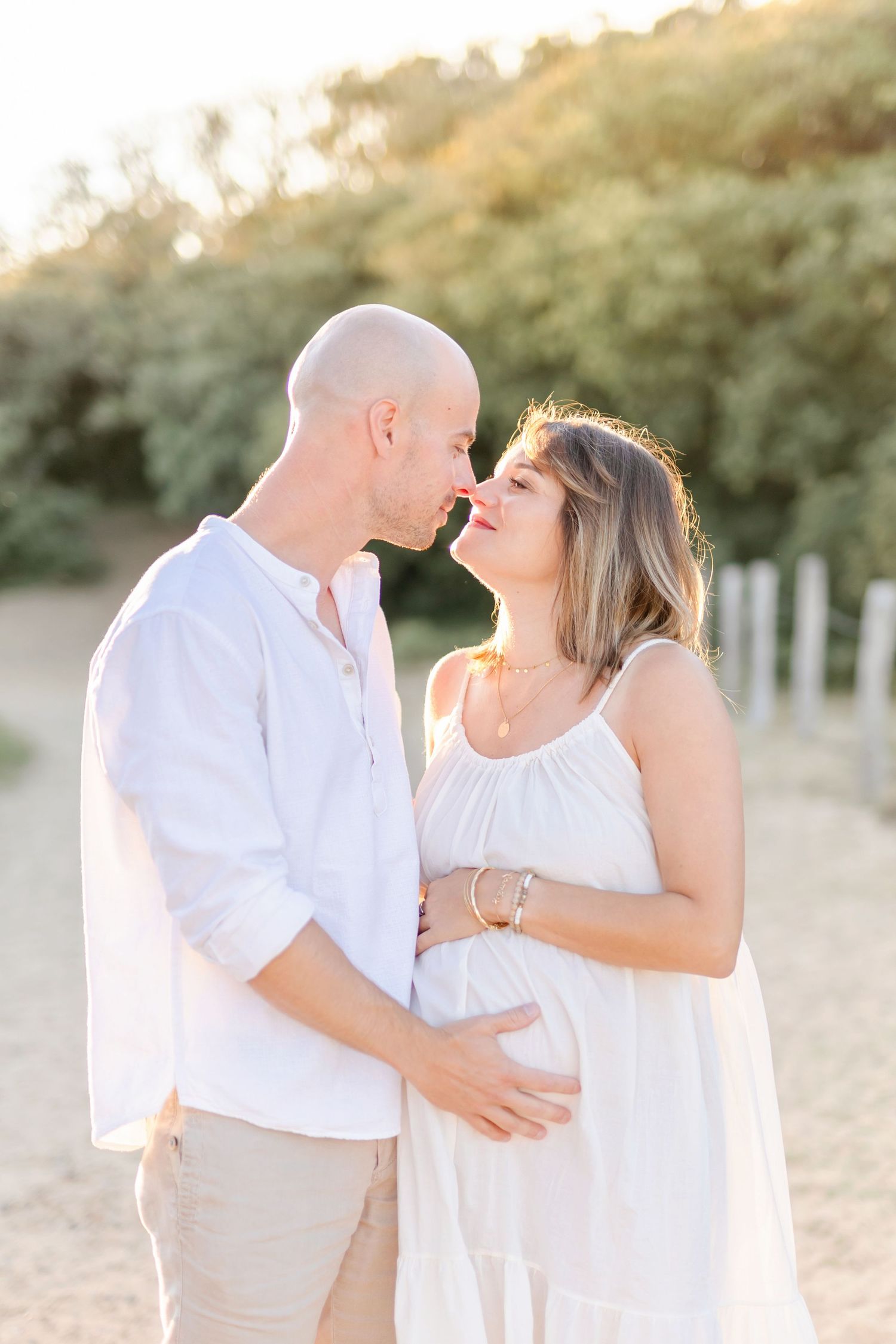 Intimate maternity portrait of couple in white sharing a tender moment on outdoor trail.