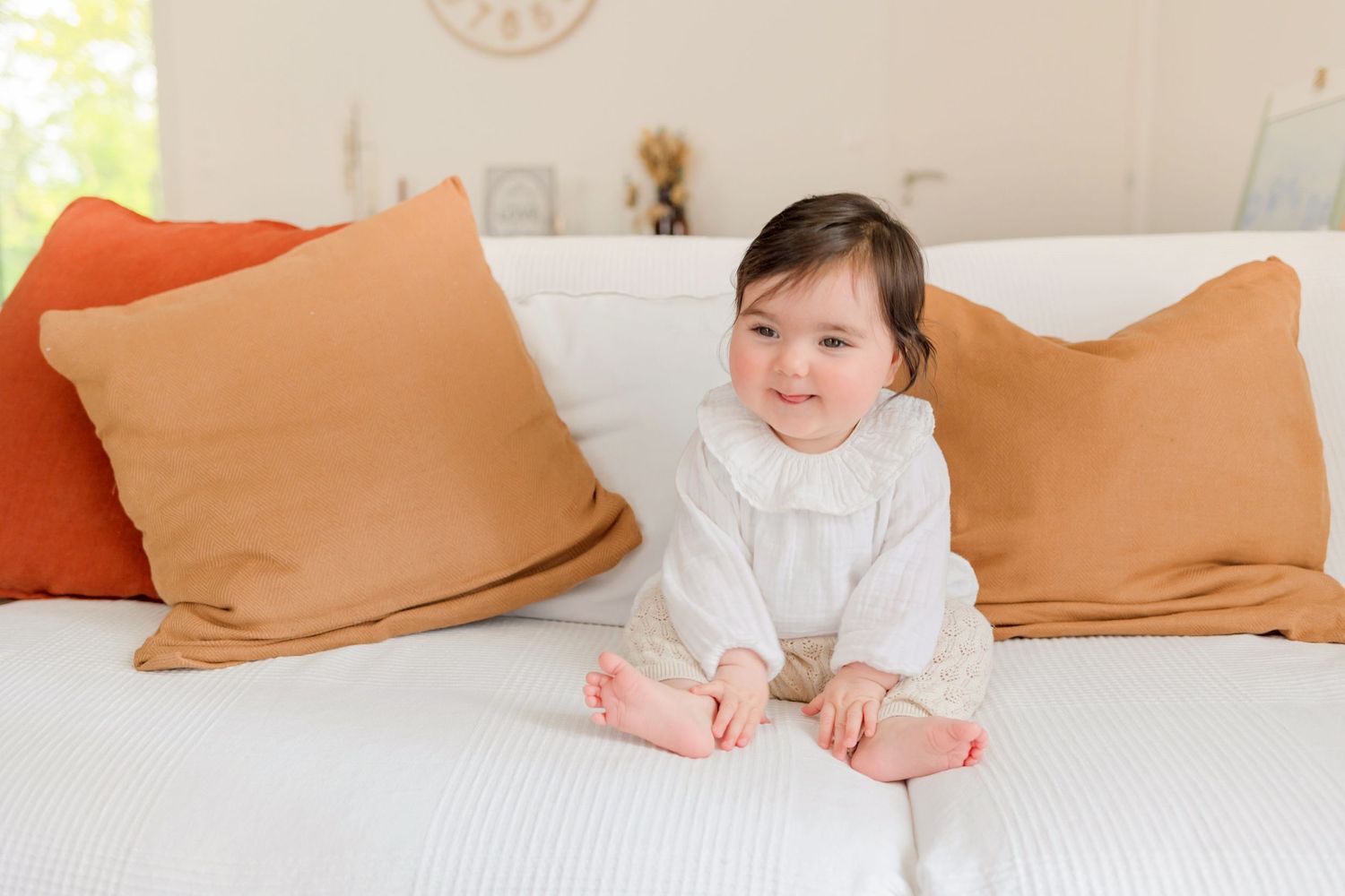 A baby in a white outfit rests on soft white bedding with orange pillows in the background.