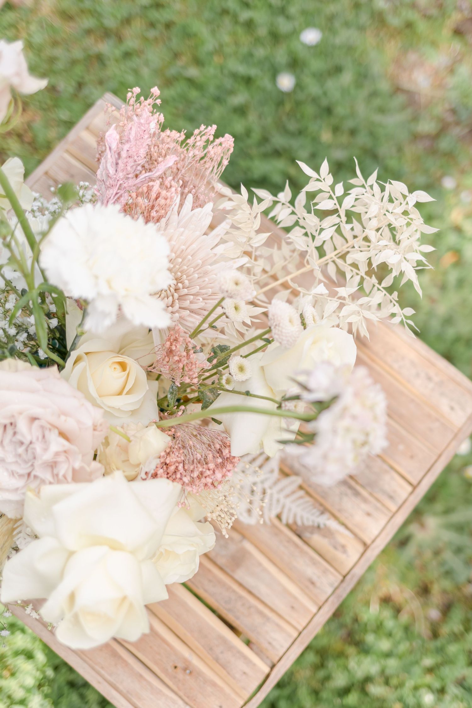 Delicate floral arrangement with white roses and pink accents displayed on wooden slats.
