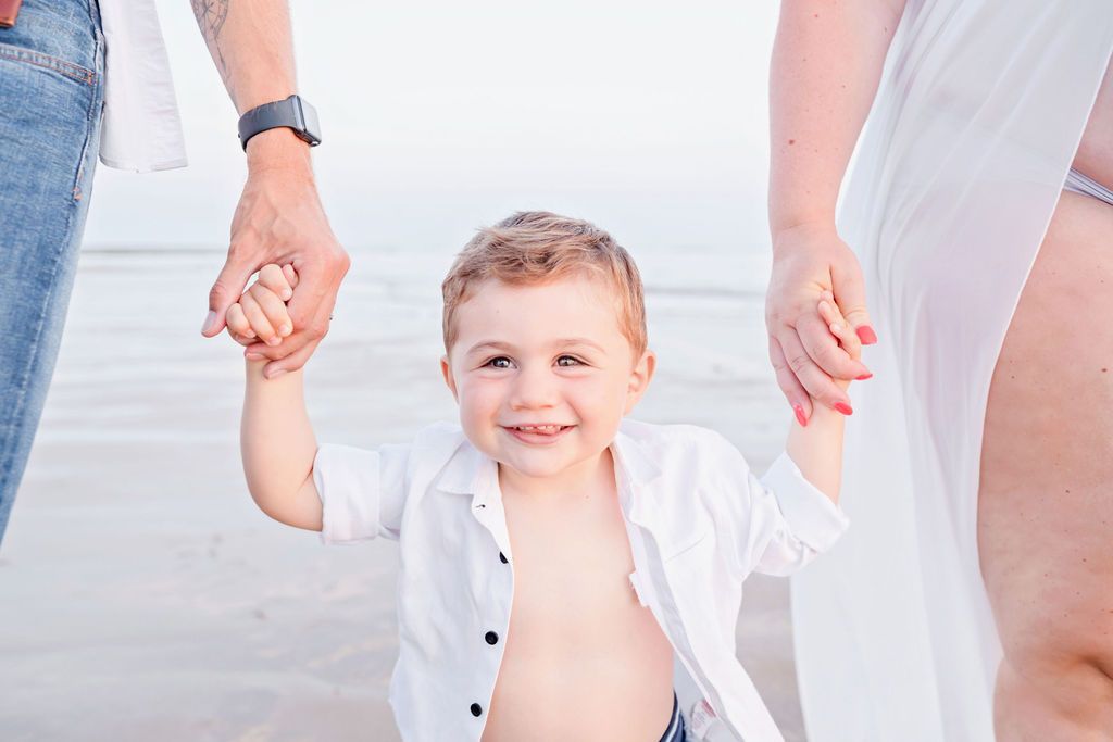 Young child holding hands of parents while walking on beach at sunset.