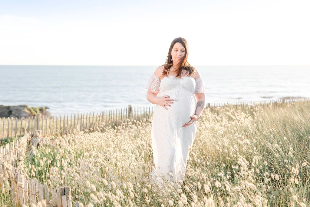 Expectant mother in white lace dress poses on coastal beach surrounded by tall grass.