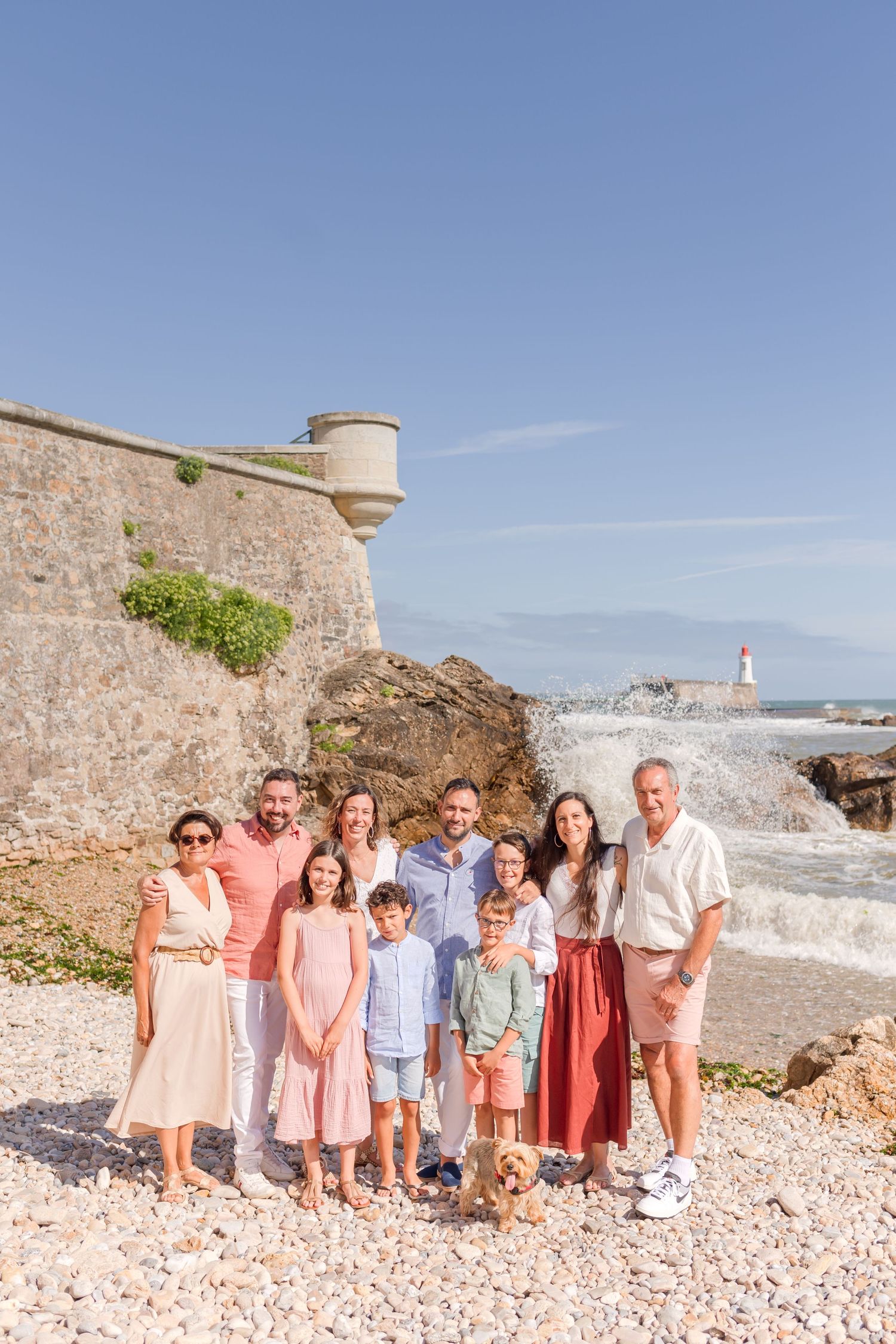 A group poses together on a rocky beach with a lighthouse and stone wall in the background on a sunny day.