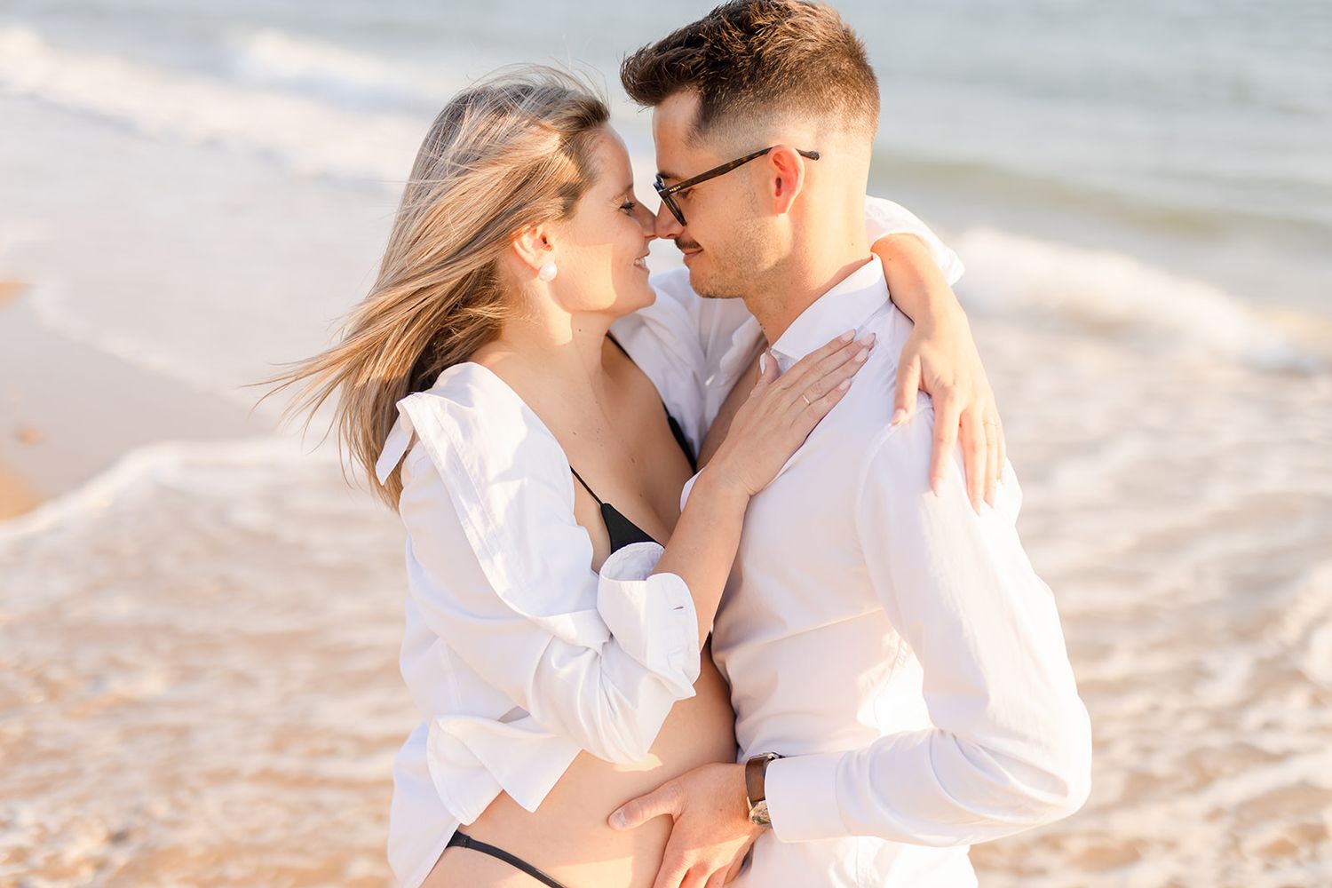 Engaged couple sharing romantic moments on a sunny beach in matching white outfits.
