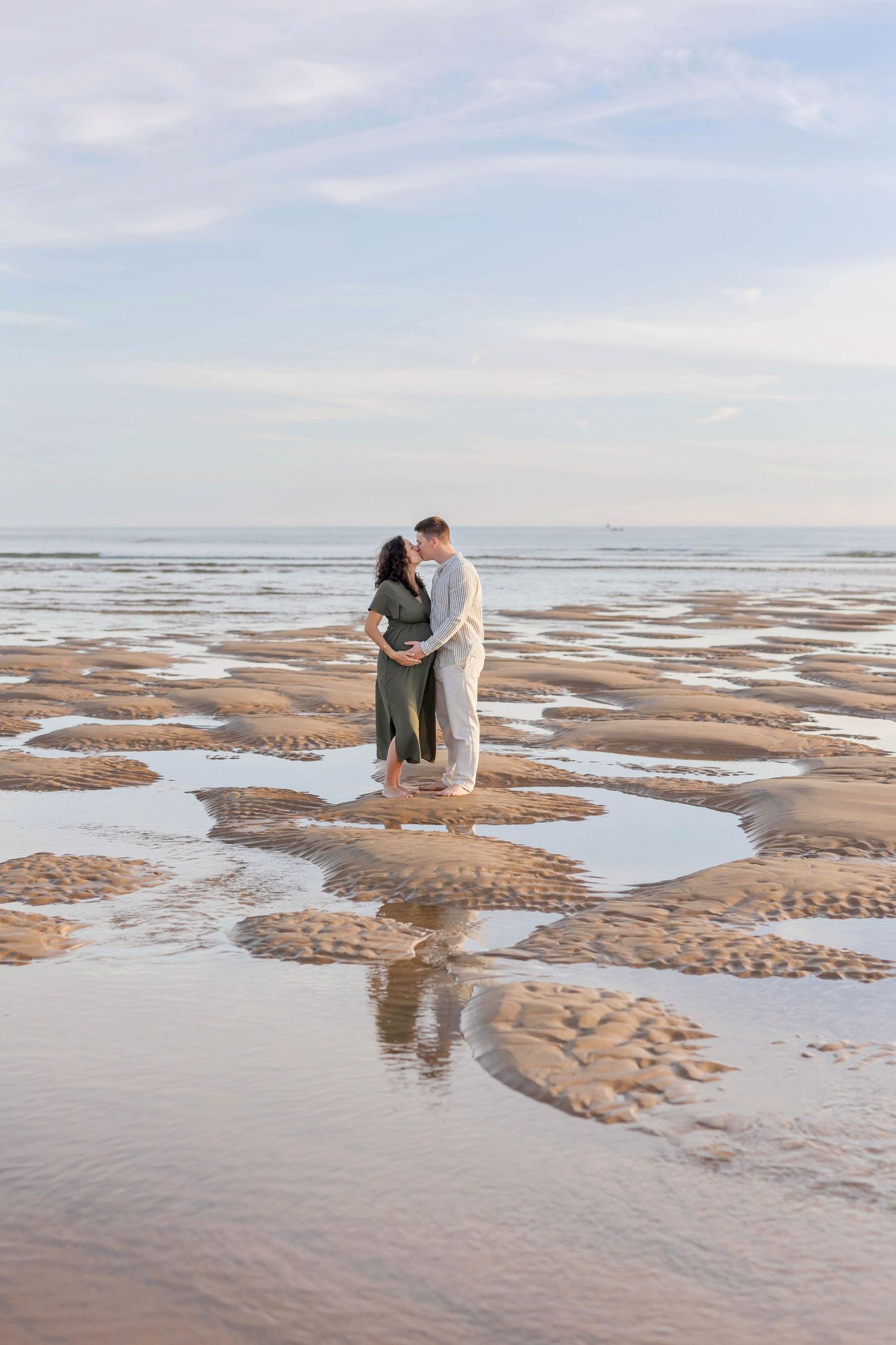 A couple embraces on a sandy beach at low tide with rippling patterns in the wet sand stretching to the horizon.