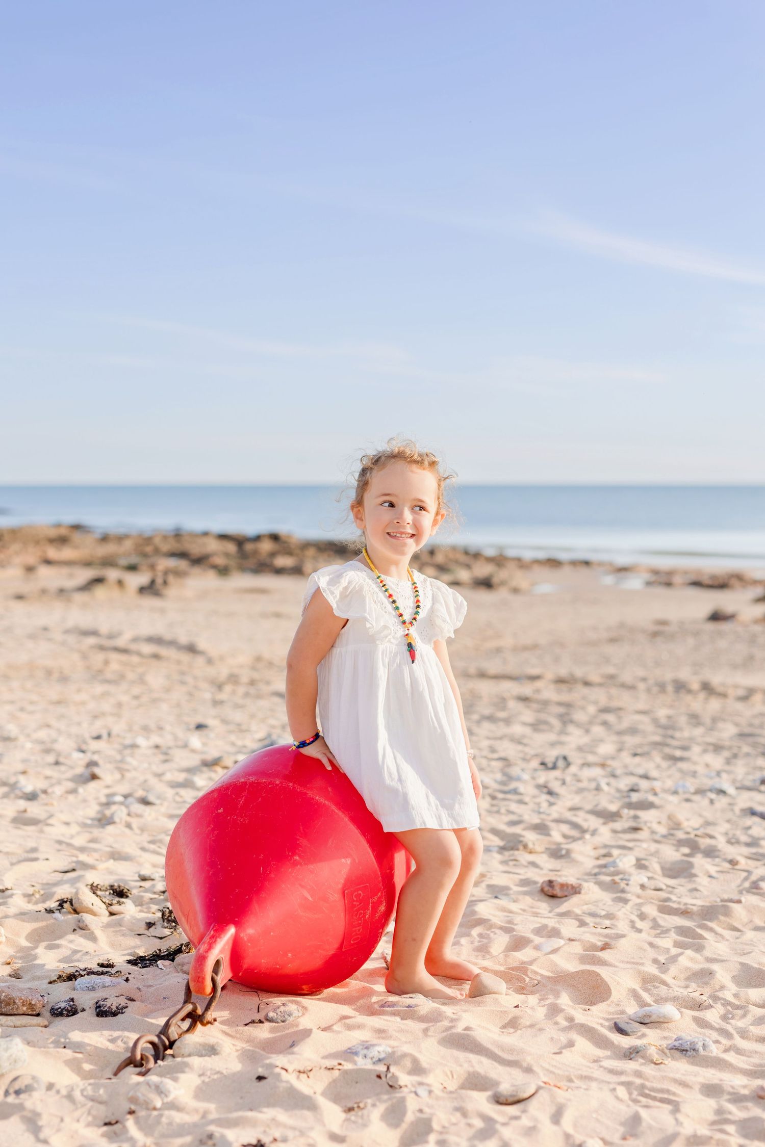 Child in white dress standing with red buoy on sunny beach with ocean in background.