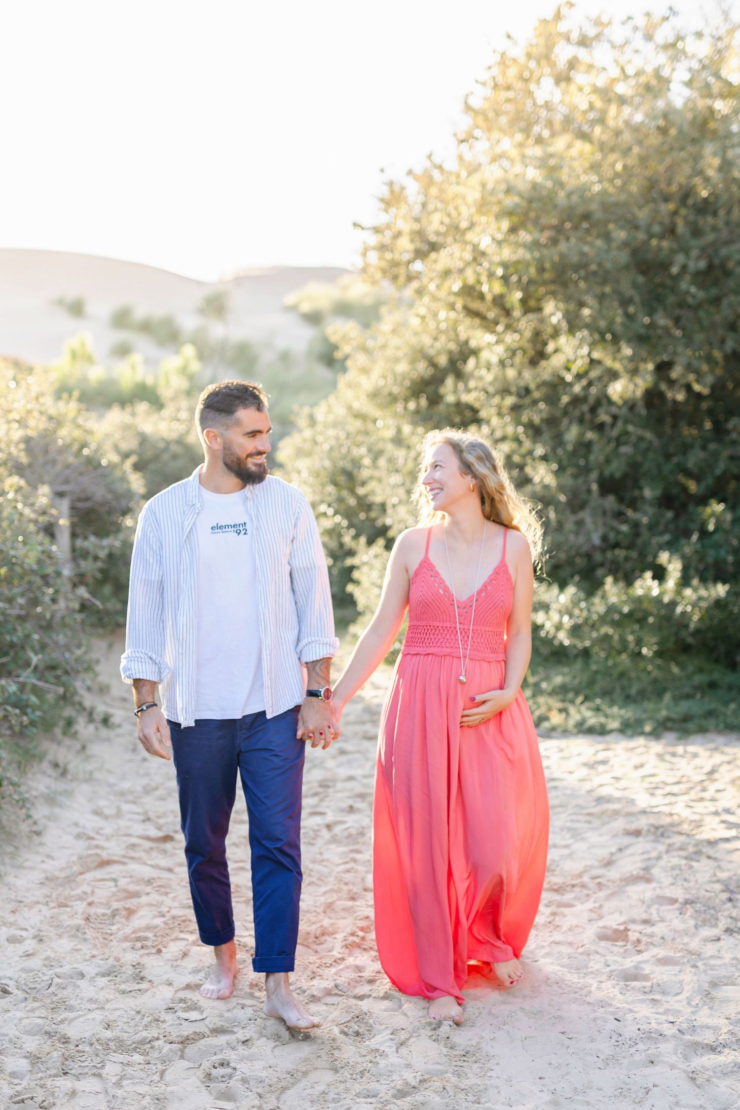 A couple walking barefoot on a sandy trail surrounded by sunlit foliage.
