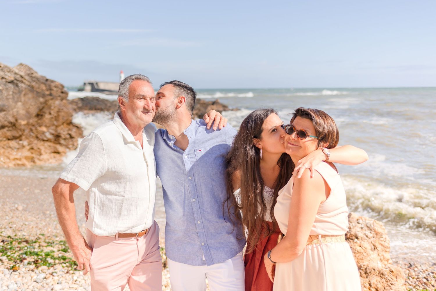 Four people share a joyful moment together on a beach with crashing waves and a lighthouse in the background.
