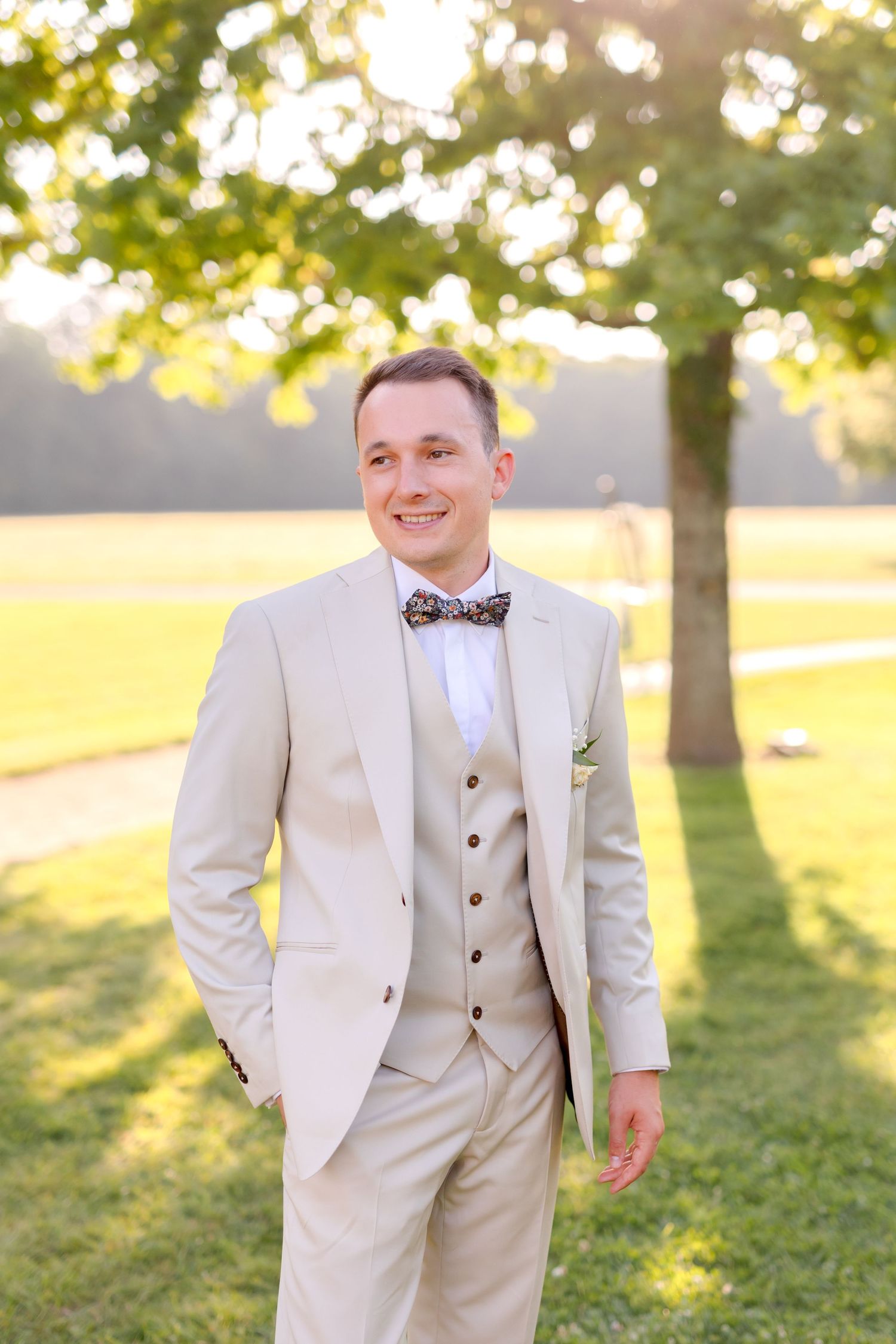 Groom in light beige three-piece suit with bowtie smiling in sunlit garden.