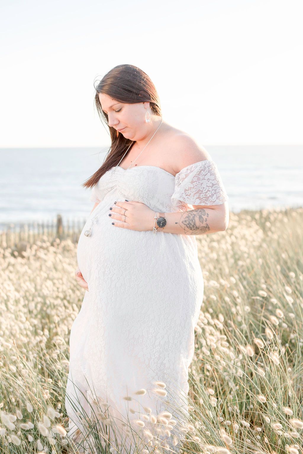 Expecting mother in off-shoulder white dress stands in coastal wildflower field.